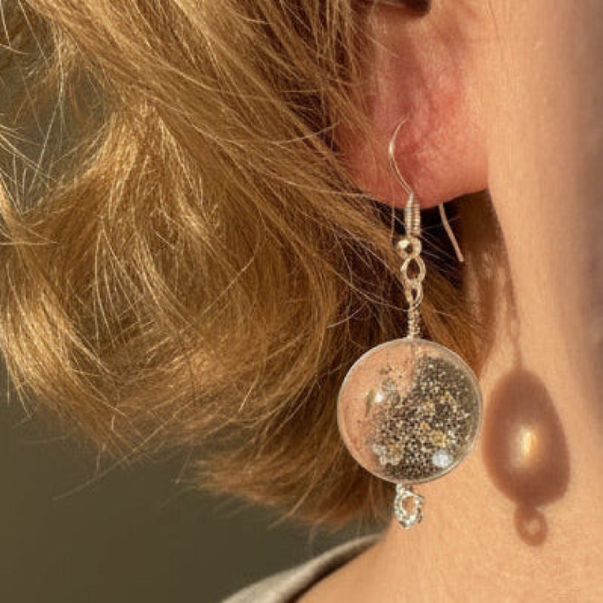 A close-up of a person wearing a dangling earring with a clear round dome filled with small gold flecks, suspended from a silver hook, with a tiny wire spiral below.