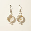 A pair of dangling earrings with clear round resin or glass pendants filled with small metallic flecks, hanging from silver fishhook wires on a brown textured background.