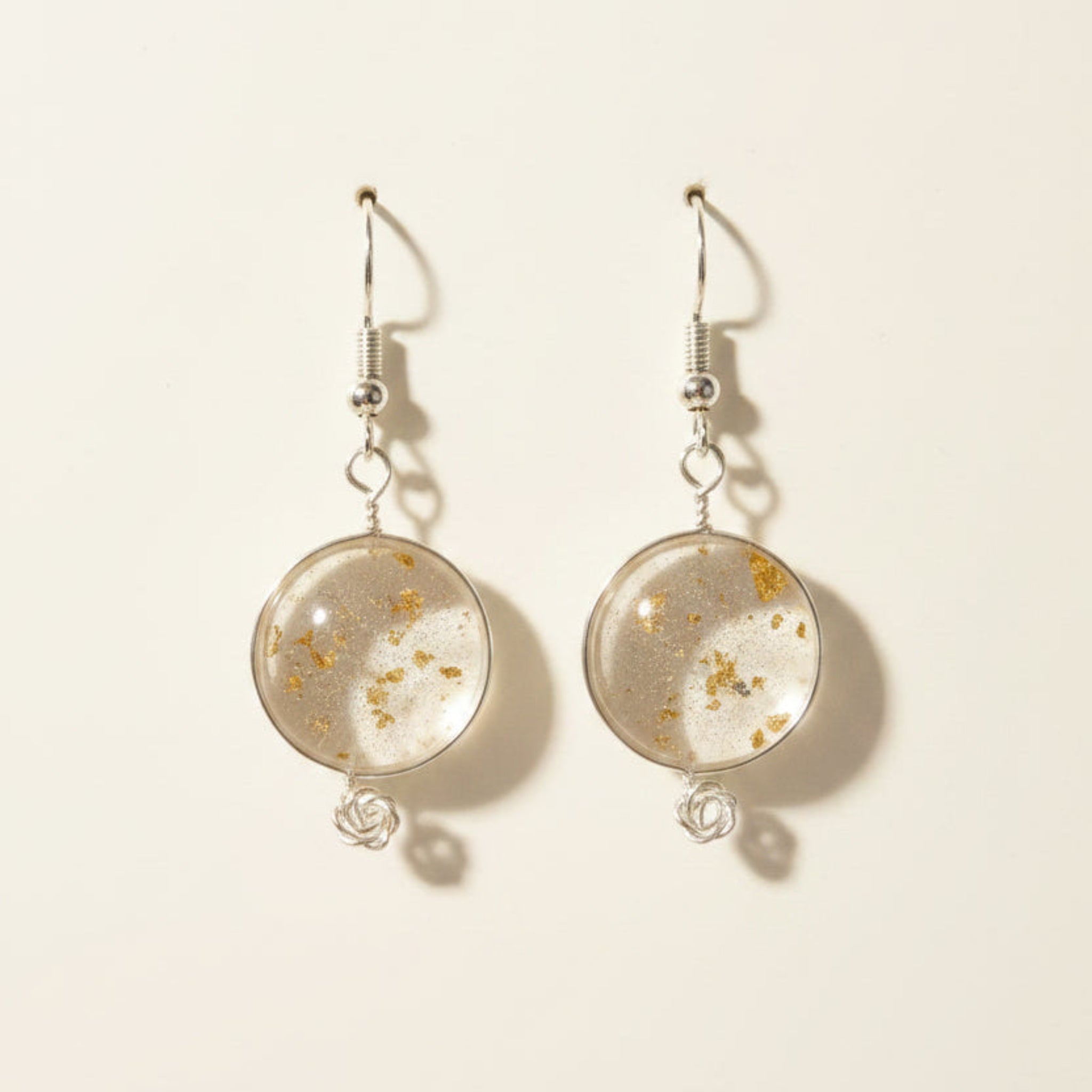 A pair of dangling earrings with clear round resin or glass pendants filled with small metallic flecks, hanging from silver fishhook wires on a brown textured background.