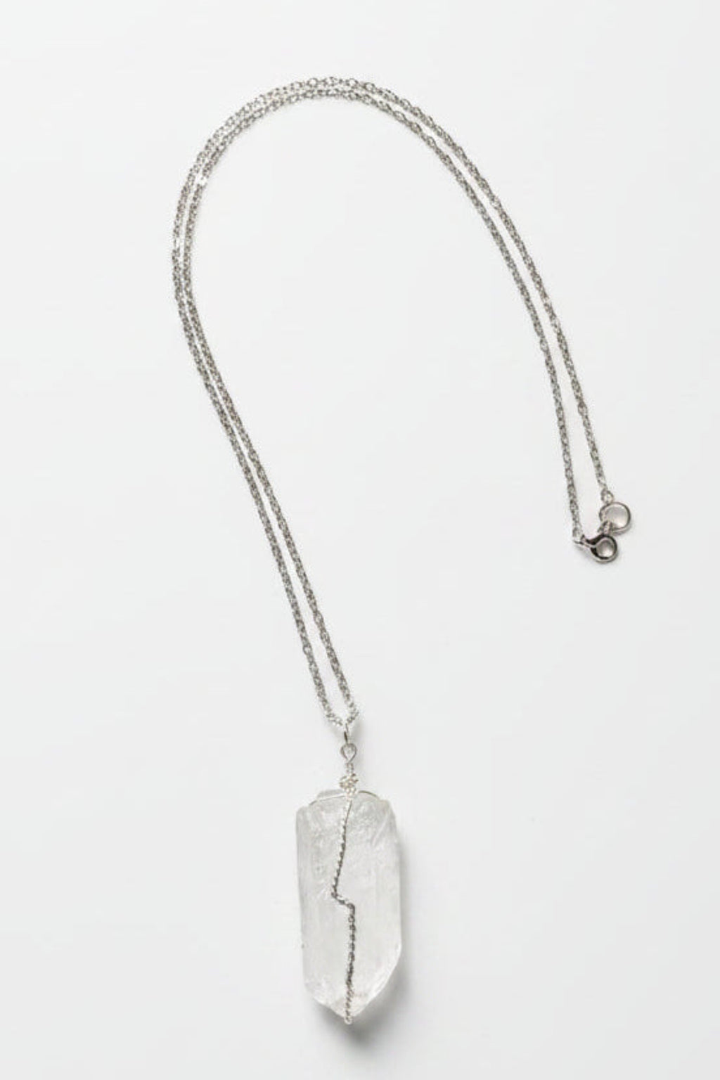 A silver-toned chain necklace with a large, irregular, clear crystal pendant, laid flat on a textured light gray fabric background.