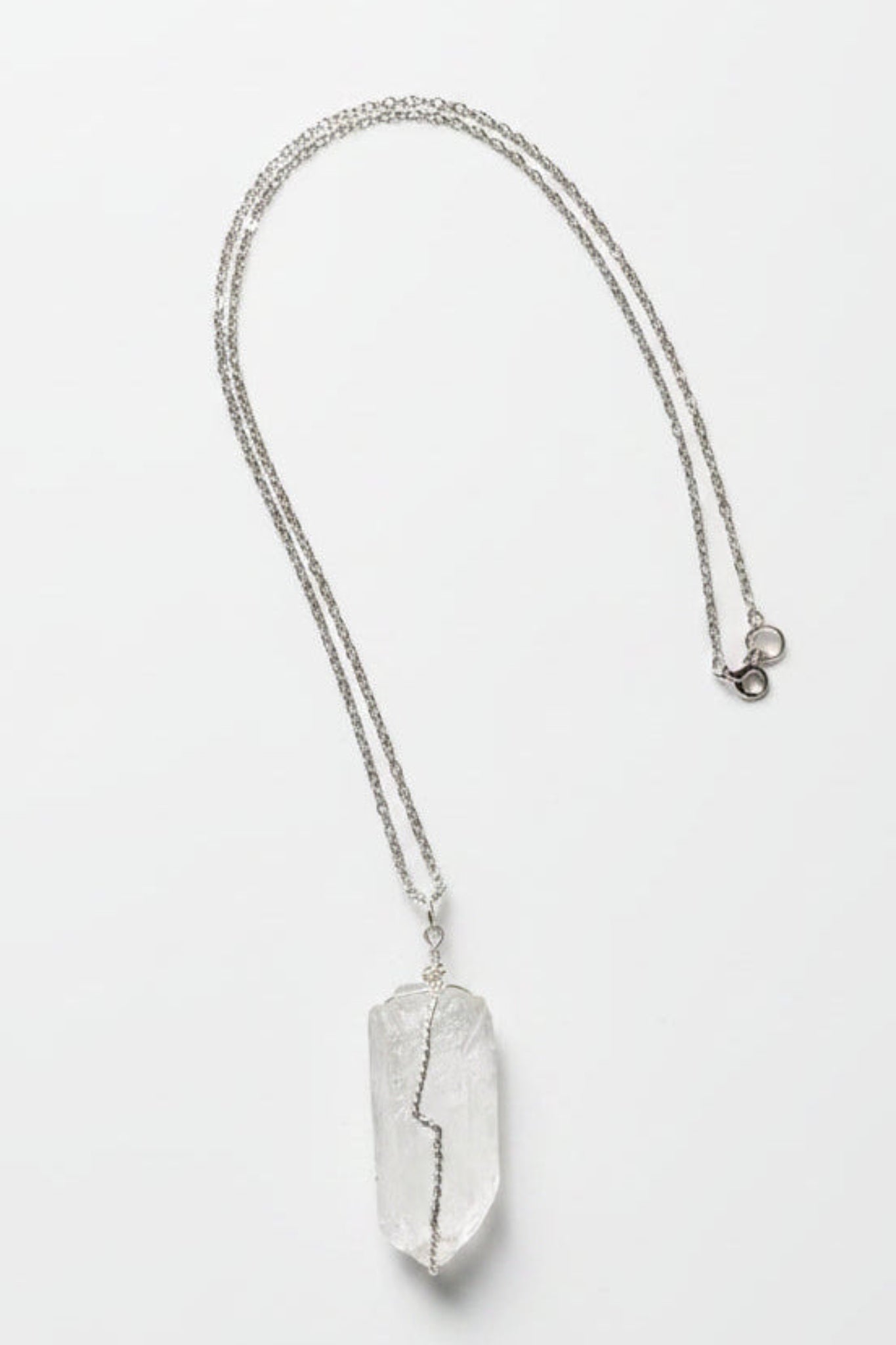 A silver-toned chain necklace with a large, irregular, clear crystal pendant, laid flat on a textured light gray fabric background.