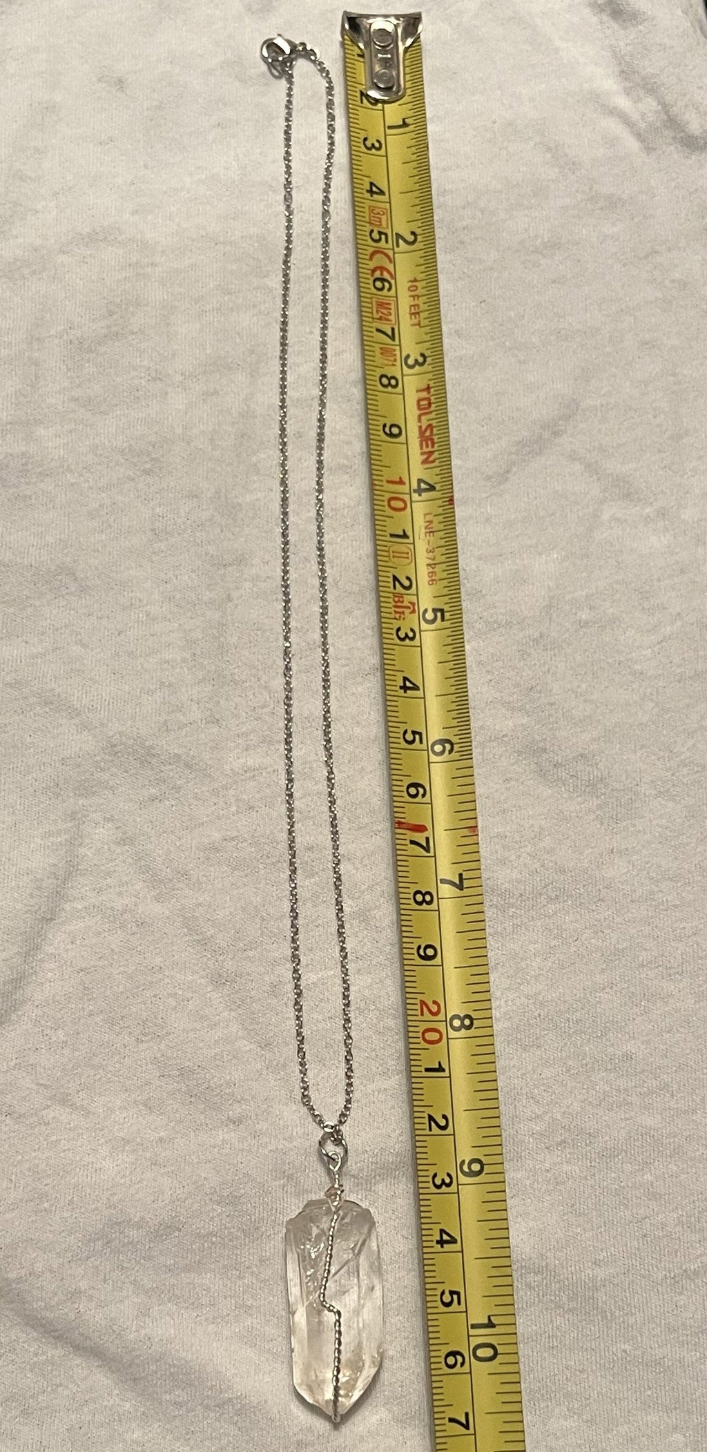 A silver chain necklace with a clear crystal pendant is shown next to a yellow tape measure on a light fabric background.