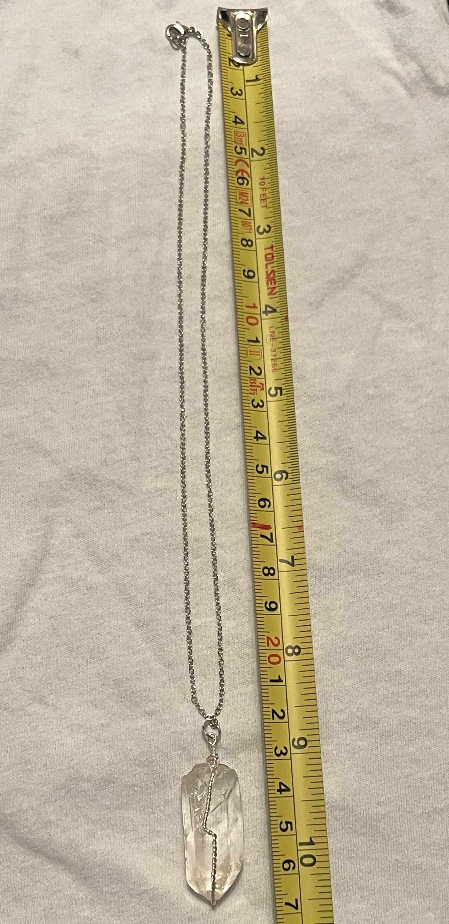 A silver chain necklace with a clear crystal pendant is shown next to a yellow tape measure on a light fabric background.