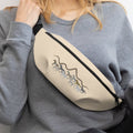 Woman sitting on the floor with a beige fanny pack with a horse and mountain design against a plain background.