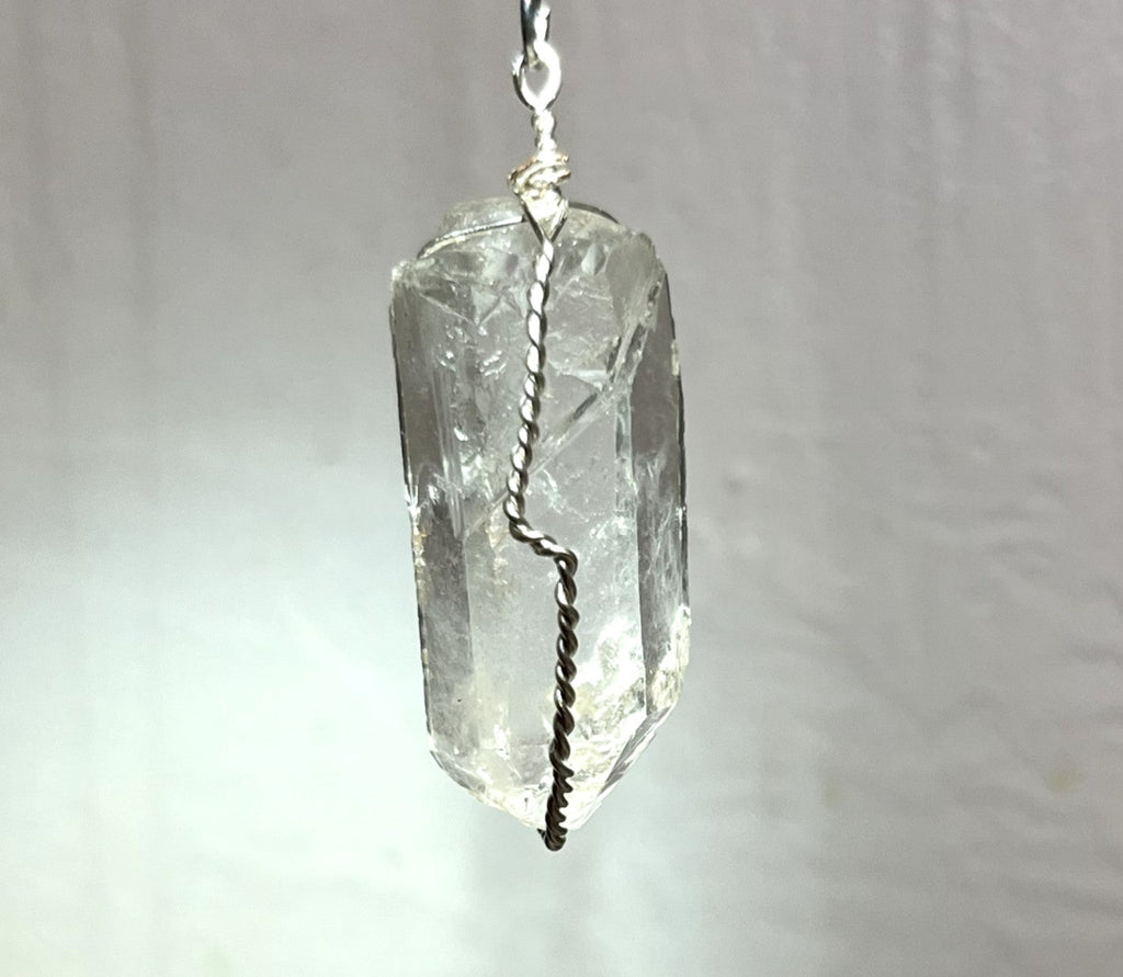A clear faceted crystal pendant, likely quartz, wrapped with a silver-colored twisted wire and a silver top loop, shown against a soft, light gray fabric background.