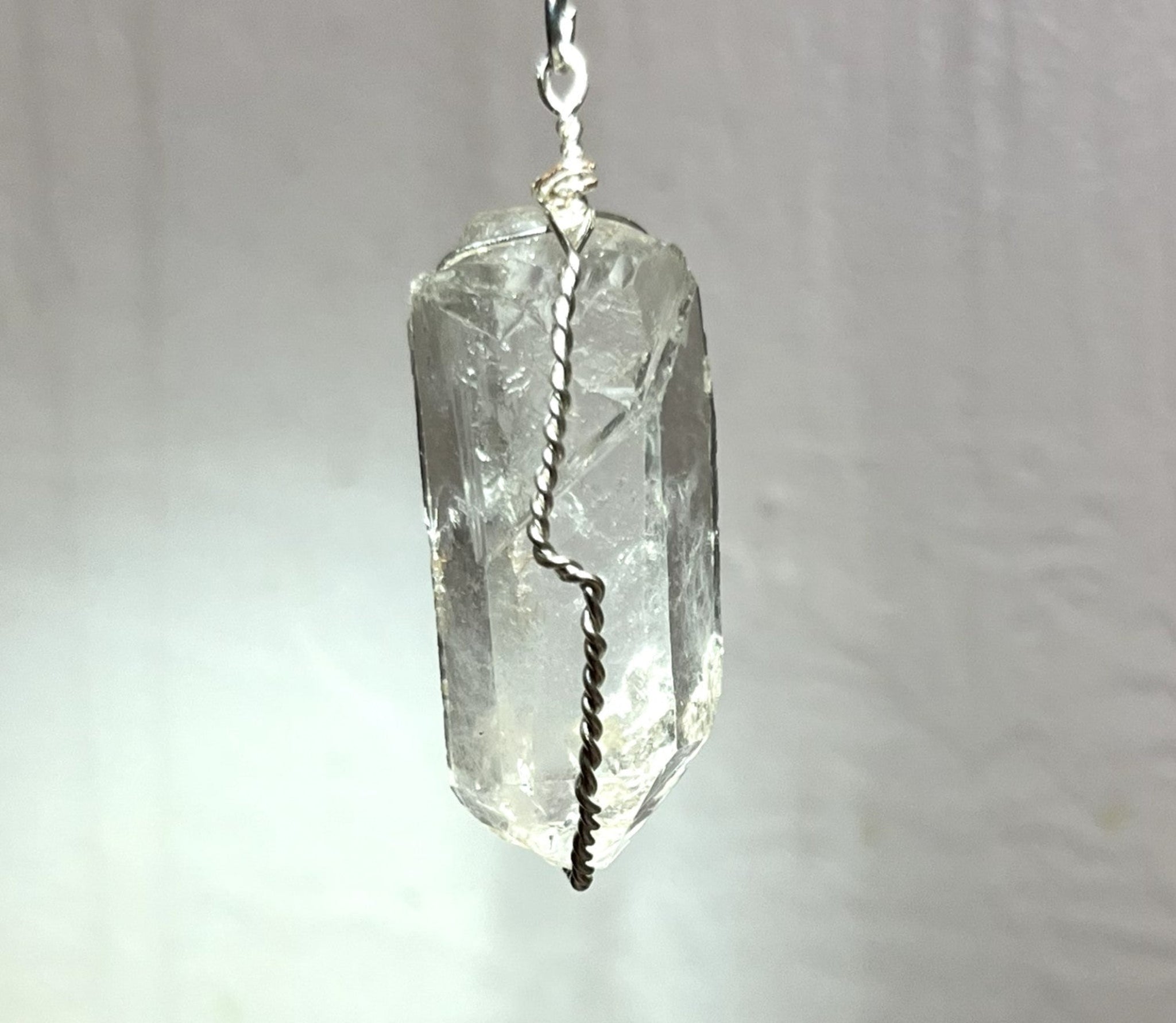 A clear faceted crystal pendant, likely quartz, wrapped with a silver-colored twisted wire and a silver top loop, shown against a soft, light gray fabric background.