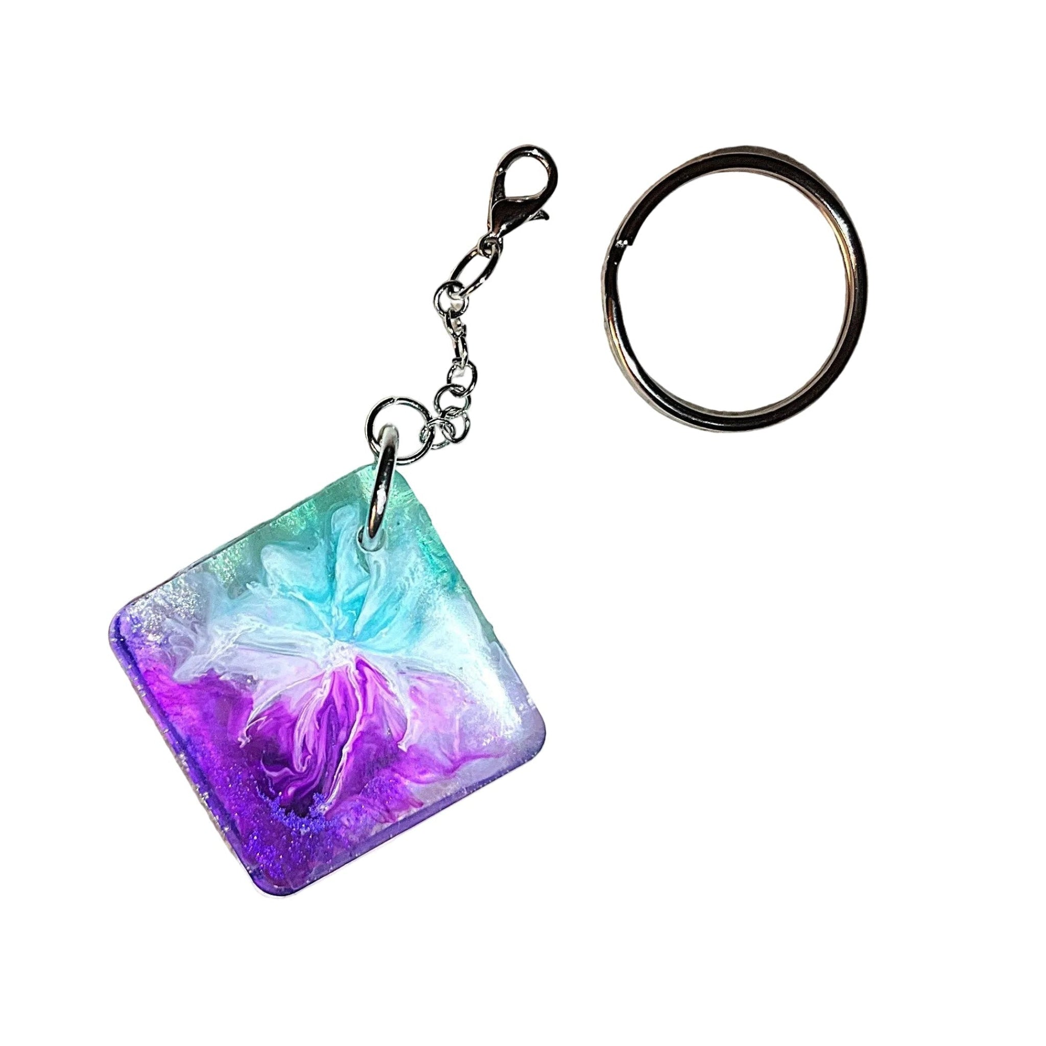 A square resin keychain charm with a colorful floral design in purple, pink, and teal, attached to a silver split keyring on a textured beige surface.