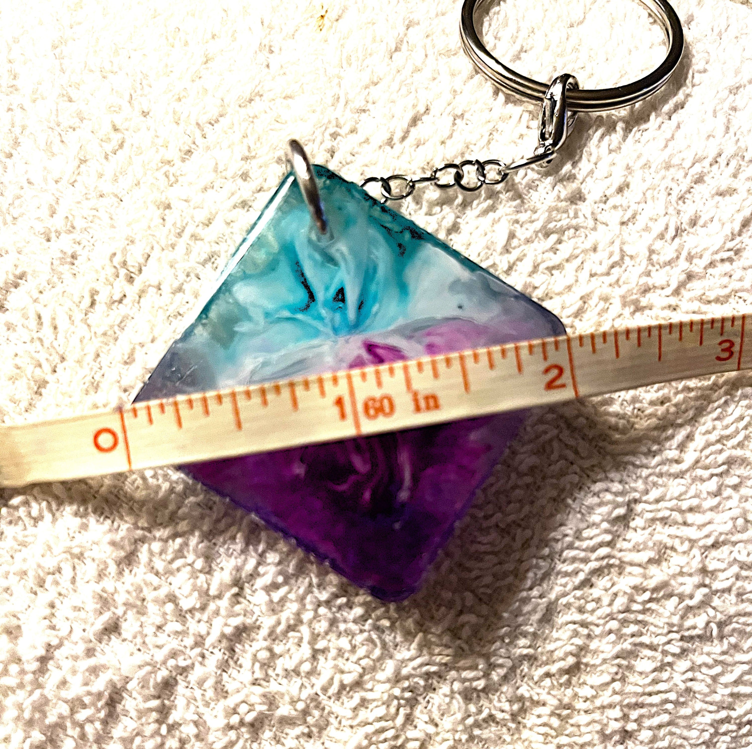 A handmade resin keychain with a diamond-shaped pendant featuring a blue, purple, and teal marbled pattern, shown with a ruler to indicate its size on a textured white fabric background.