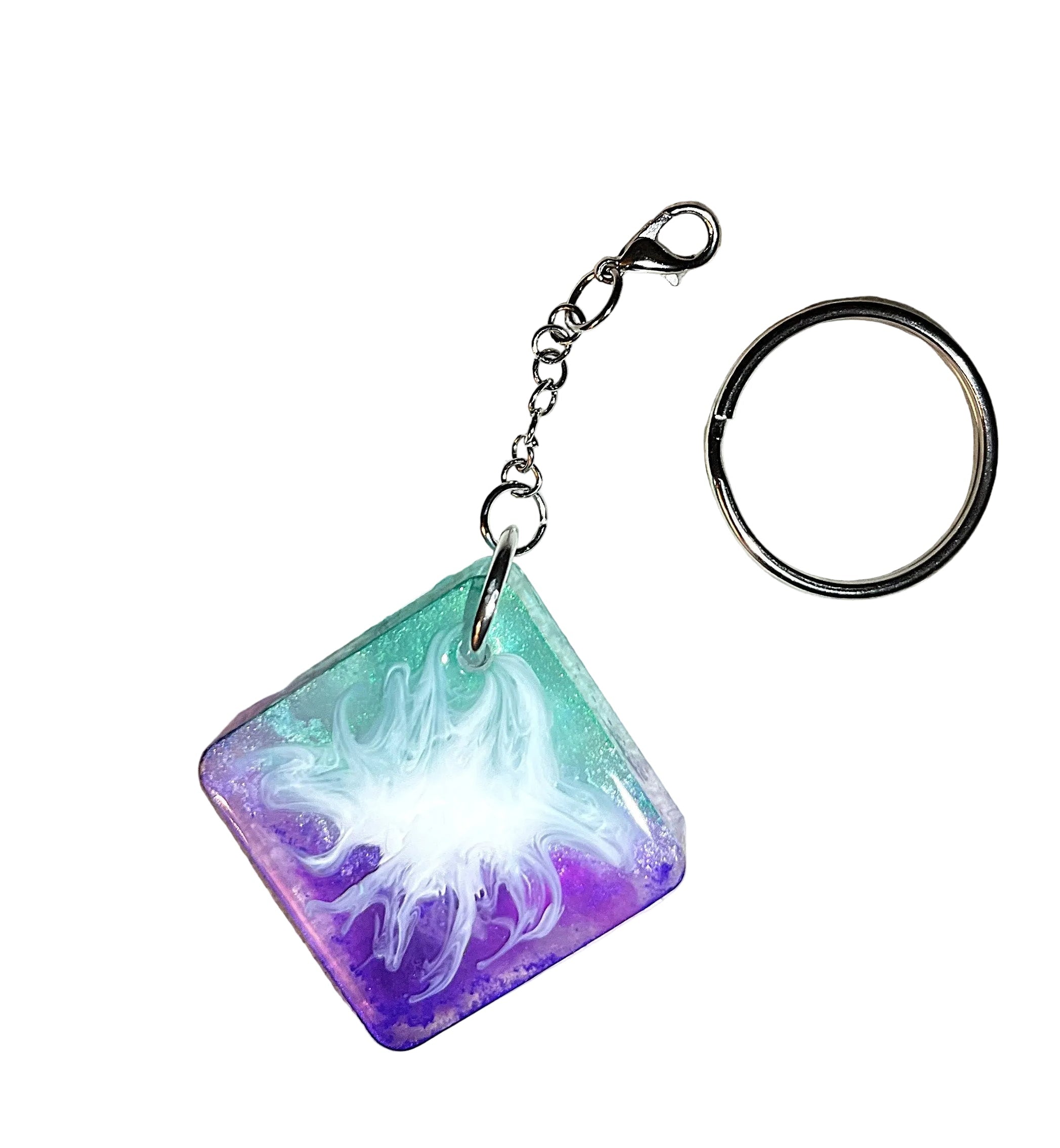 A square resin keychain with a silver split ring and clasp, featuring a colorful design of teal, purple, and white swirling patterns, resting on a textured beige surface.