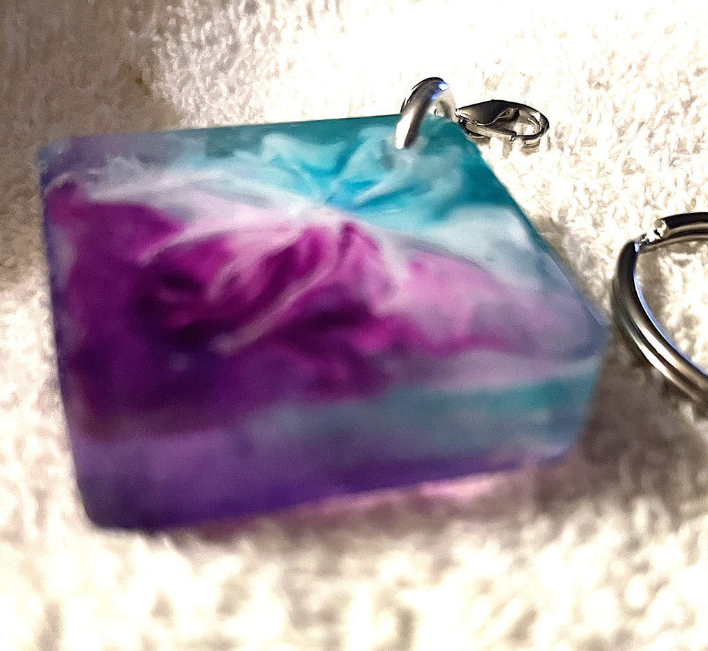 A square resin pendant keychain with a marbled swirl pattern of purple, teal blue, and white, featuring a silver lobster clasp and key ring on a textured white fabric background.