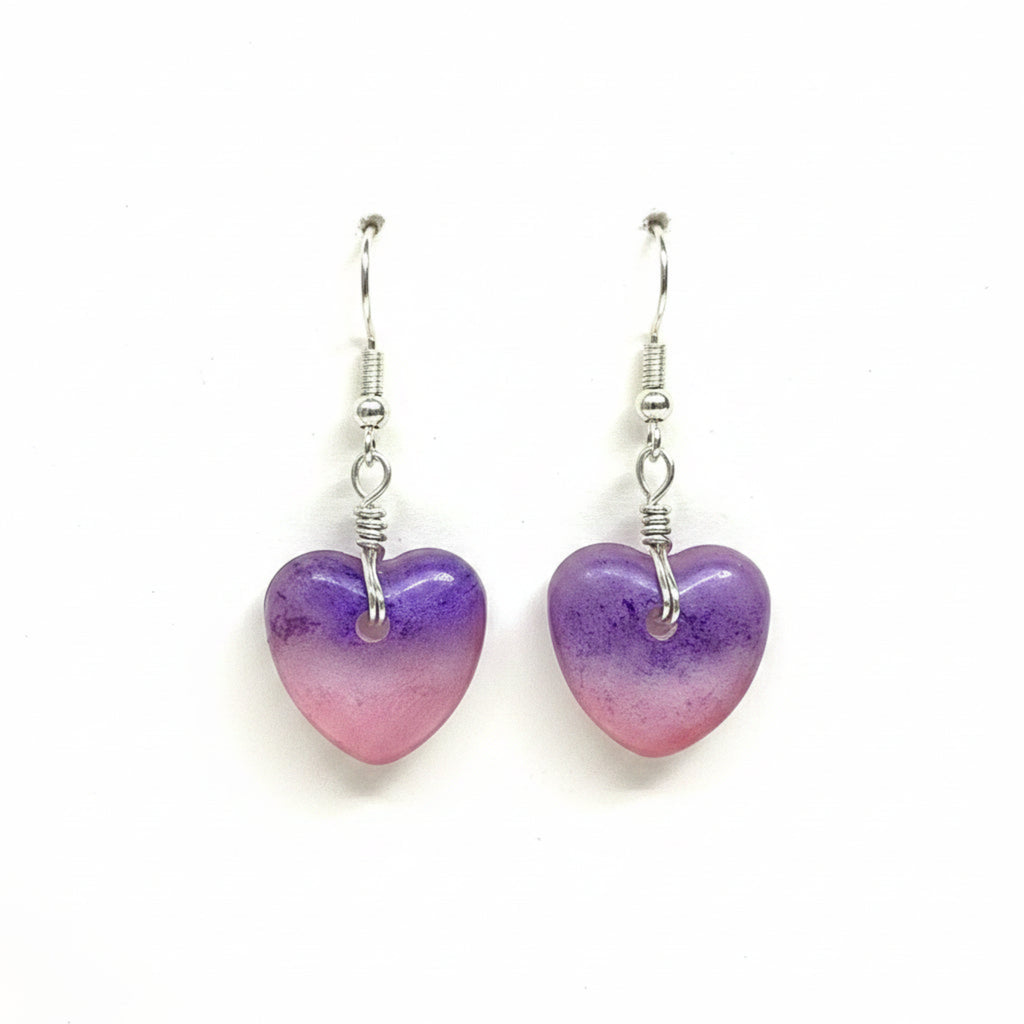 A pair of heart-shaped dangle earrings made from translucent purple and pink resin, suspended from silver fishhook wires on a plain white background.