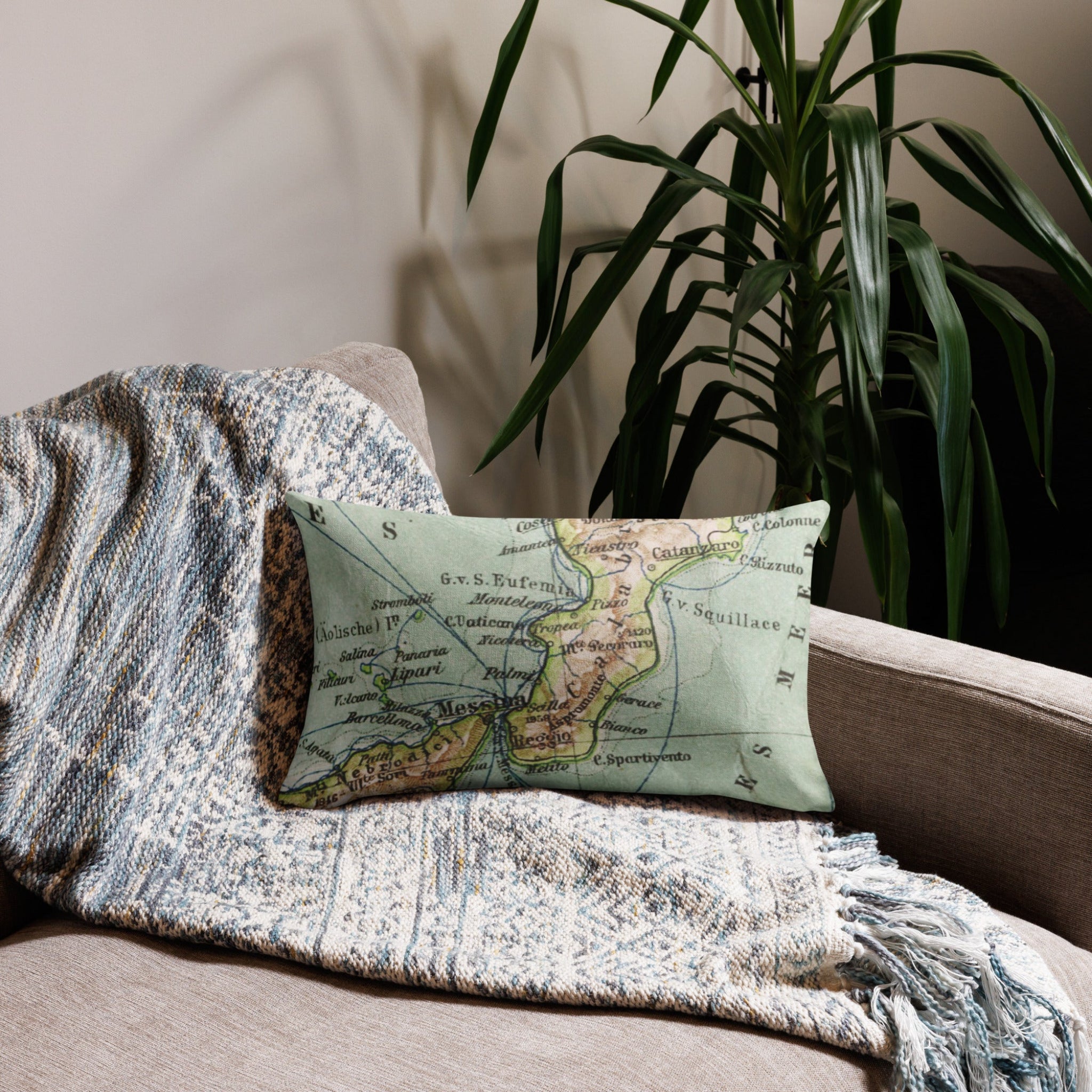Decorative pillow with a map pattern on a beige sofa with a textured blanket and plant in the background.