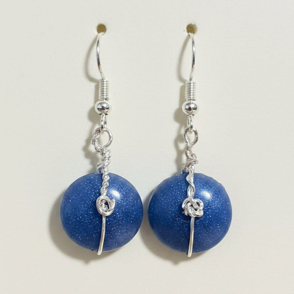 A pair of handmade dangle earrings featuring round, glittery blue beads suspended from silver hooks with twisted wire details, displayed on a brown textured surface.