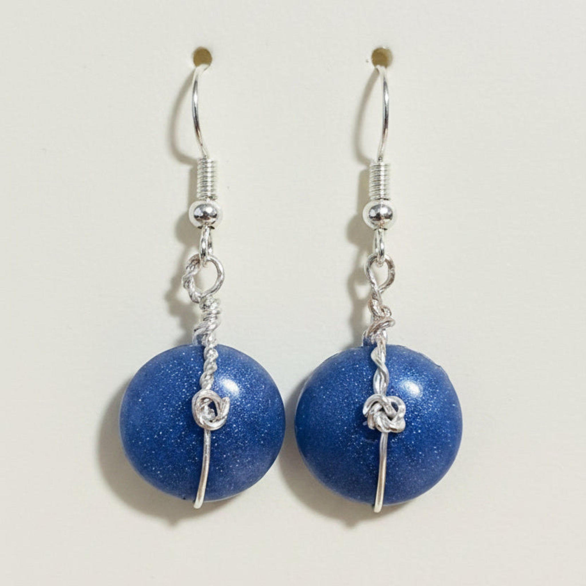 A pair of handmade dangle earrings featuring round, glittery blue beads suspended from silver hooks with twisted wire details, displayed on a brown textured surface.