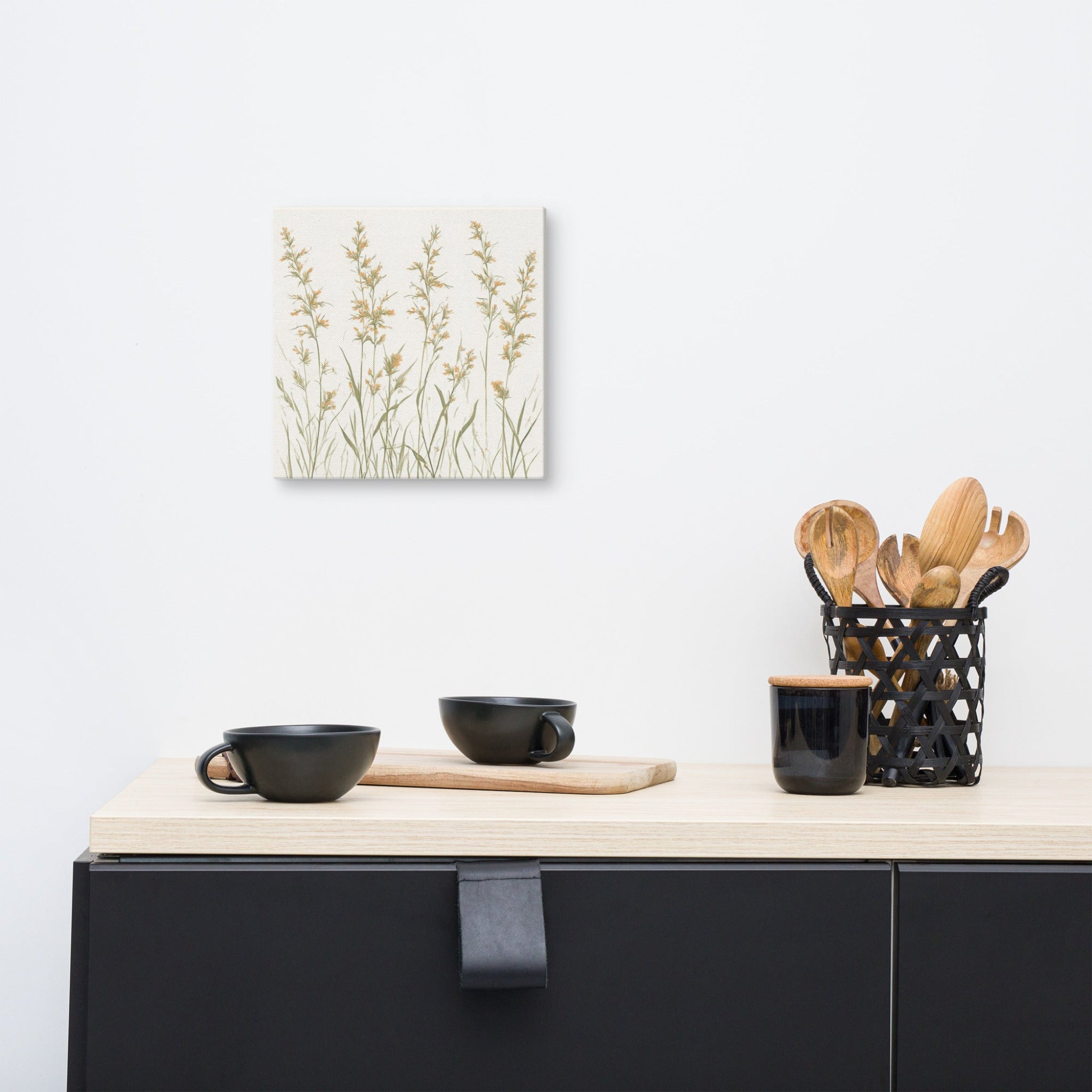 A square canvas print featuring minimalist illustrations of delicate, neutral-toned meadow grasses and wildflower stems on a cream background. Kitchen counter with black bowls, a cutting board, and wooden utensils on a light background.