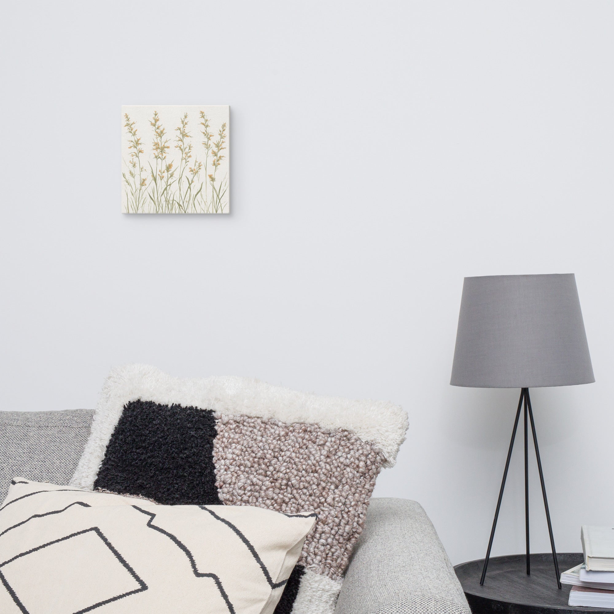 A square canvas print featuring minimalist illustrations of delicate, neutral-toned meadow grasses and wildflower stems on a cream background and Decorative pillows on a sofa with a lamp in the background.