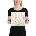 A woman holding a square canvas print featuring minimalist illustrations of delicate, neutral-toned meadow grasses and wildflower stems on a cream background.