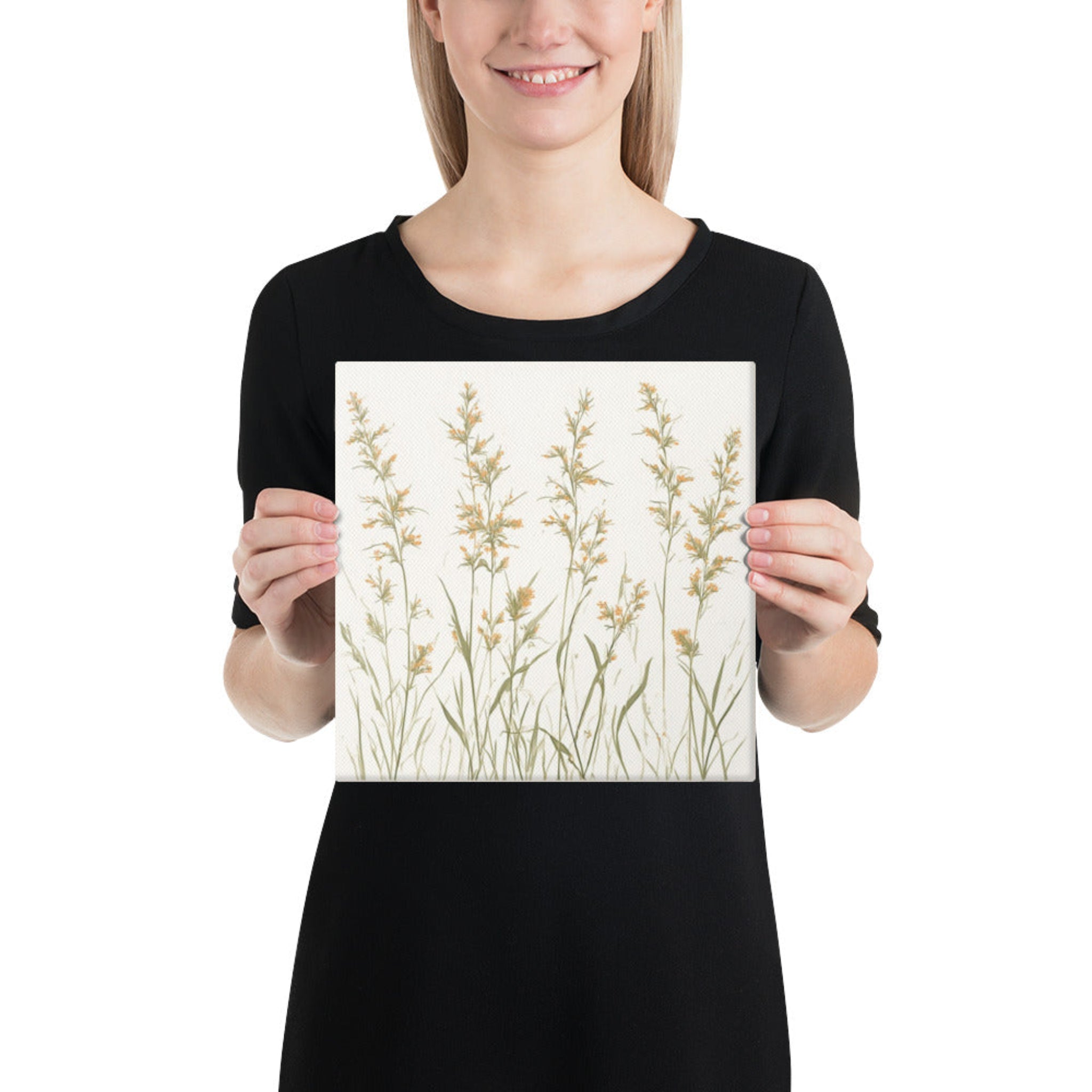 A woman holding a square canvas print featuring minimalist illustrations of delicate, neutral-toned meadow grasses and wildflower stems on a cream background.