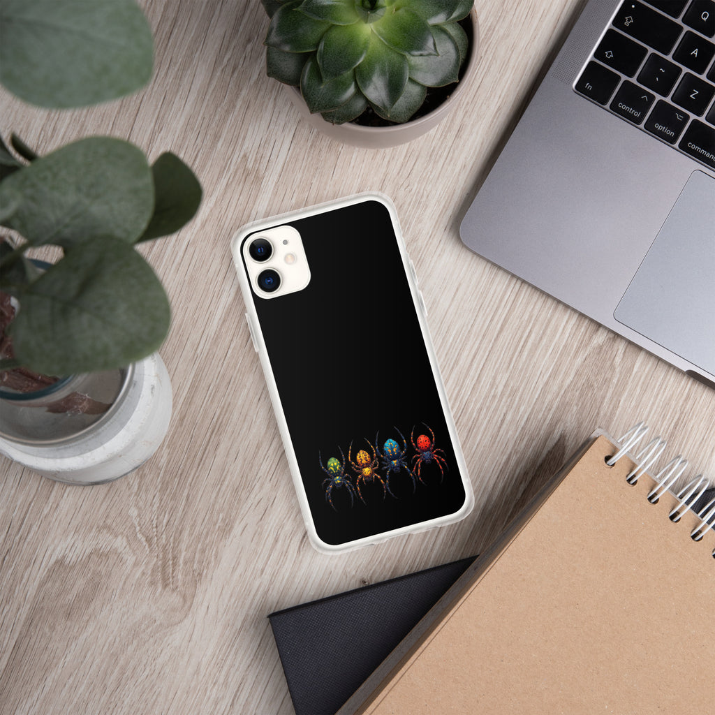 A smartphone case with a black back featuring a graphic of four colorful spiders, shown in a styled flat-lay on a light wood desk next to a laptop, notebook, and plants.