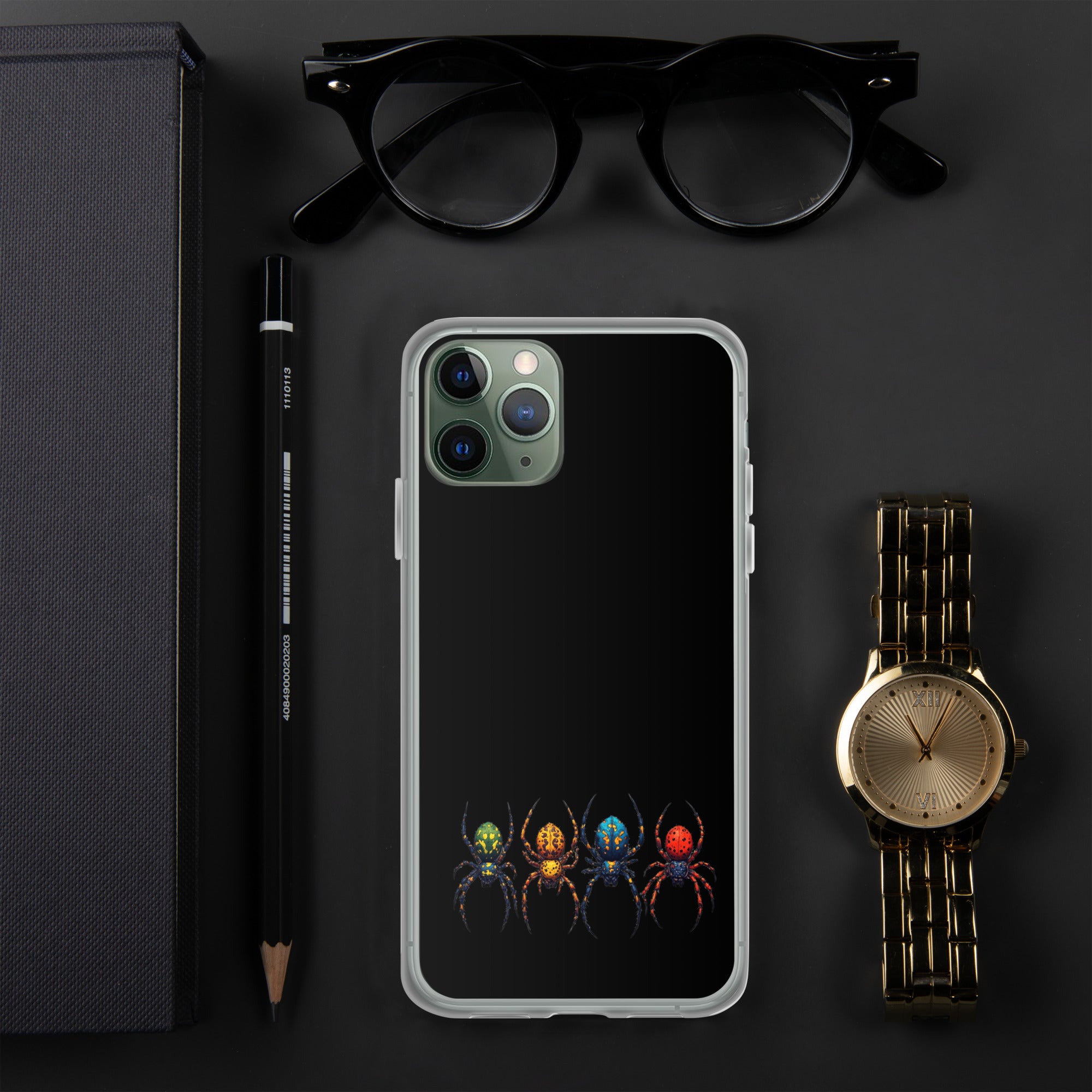 A clear iPhone case with a black back panel featuring a graphic of four colorful spiders at the bottom, styled in a flat-lay with black eyeglasses, a black pencil, a black notebook, and a gold-tone wristwatch on a dark gray background.