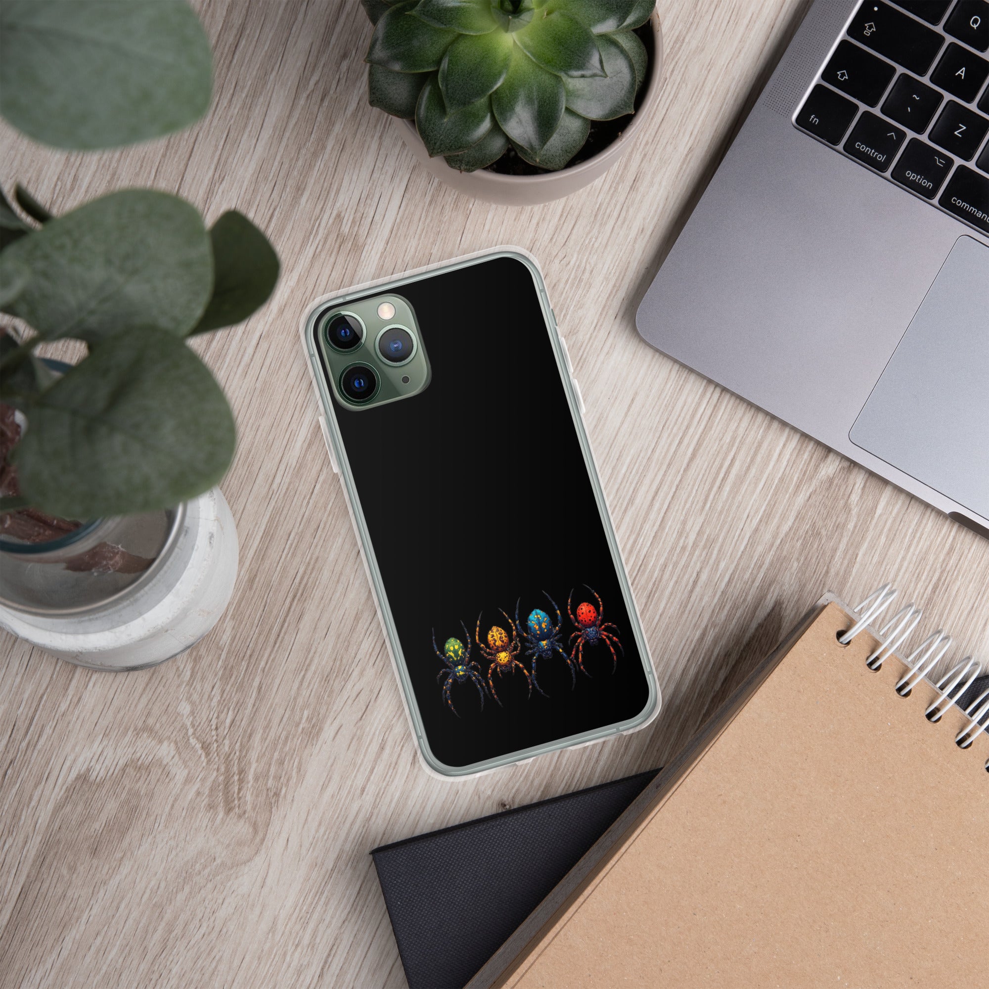 A black phone case with a graphic of four colorful spiders at the bottom, shown on a light wood desk with a laptop, plants, and a notebook.