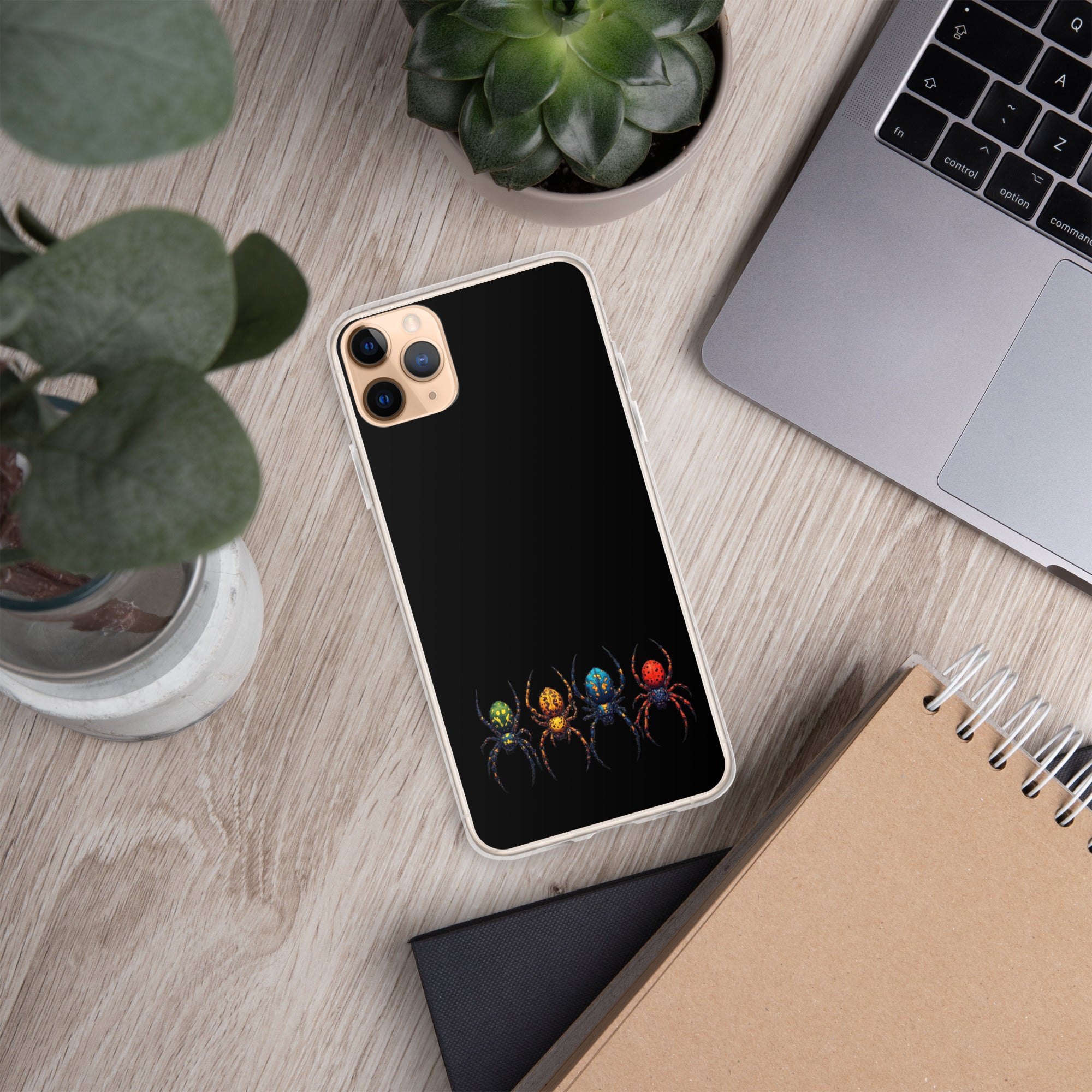 A black phone case with a colorful spider design, shown on a silver smartphone, placed on a light wood desk with a laptop, plants, and a notebook nearby.