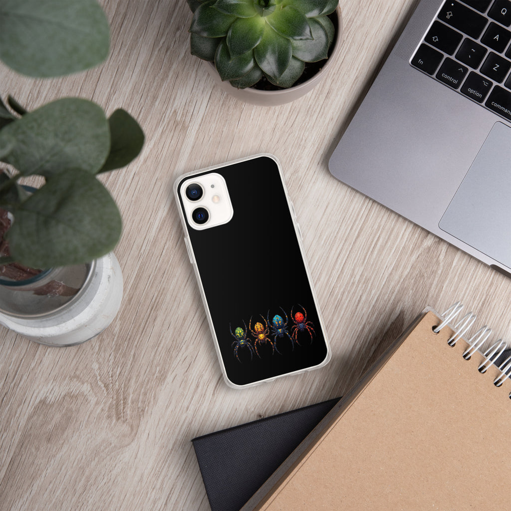 A black phone case with a graphic of four colorful spiders at the bottom, shown on a light wood desk with a laptop, plants, and notebooks.