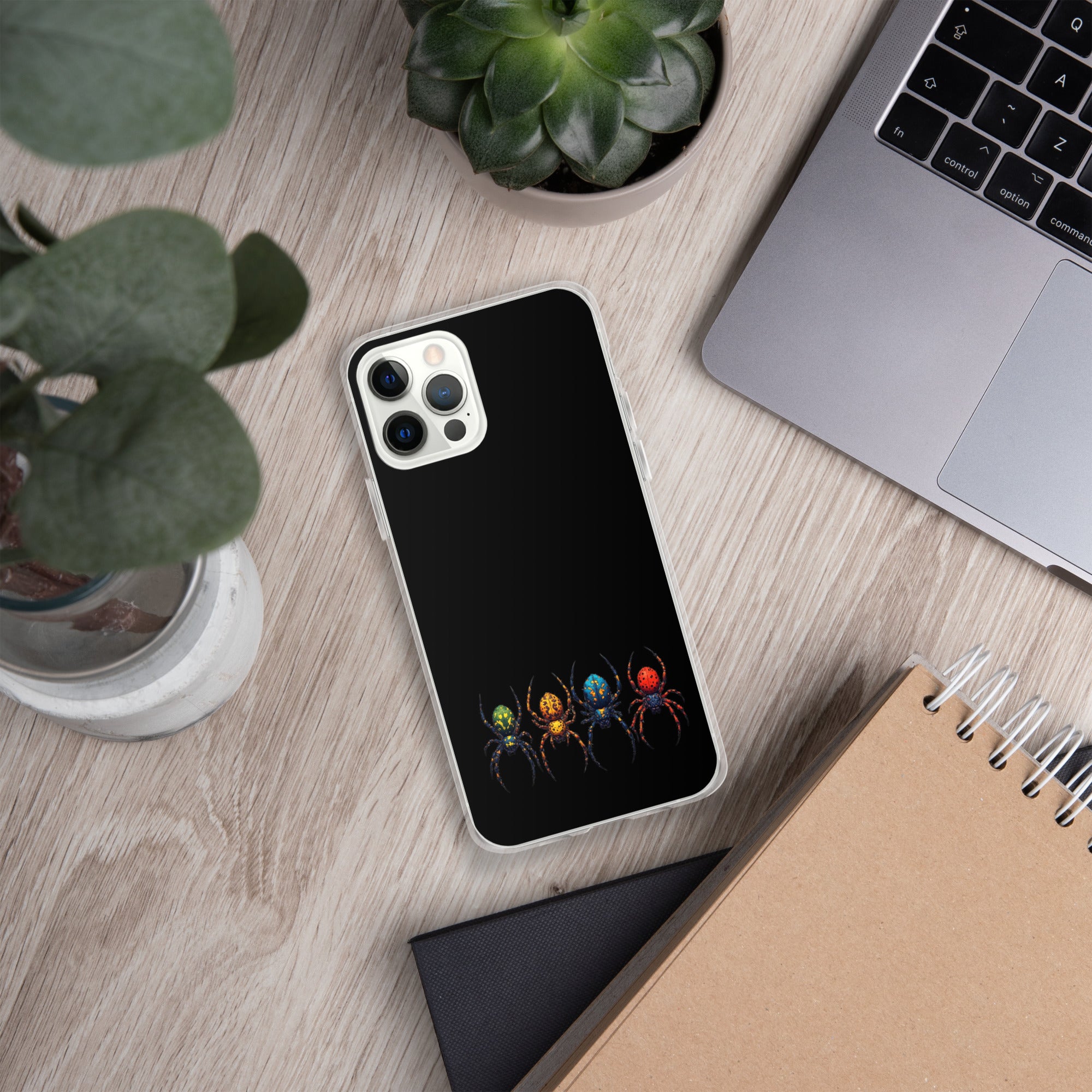 A black phone case with a graphic of four colorful spiders at the bottom, shown on a silver smartphone in a styled flat-lay desk setting with a laptop, plants, and a notebook.