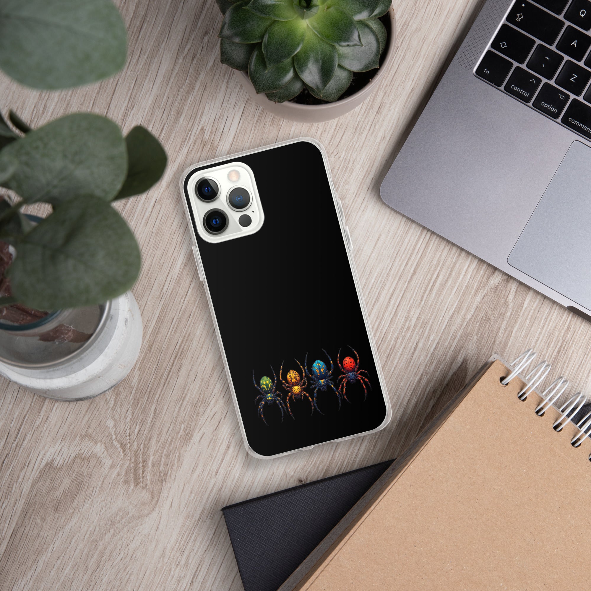A black phone case with a graphic of four colorful spiders, shown on a silver smartphone. The phone case is displayed in a styled flat-lay setting on a light wood desk alongside a laptop, plants, and notebooks.