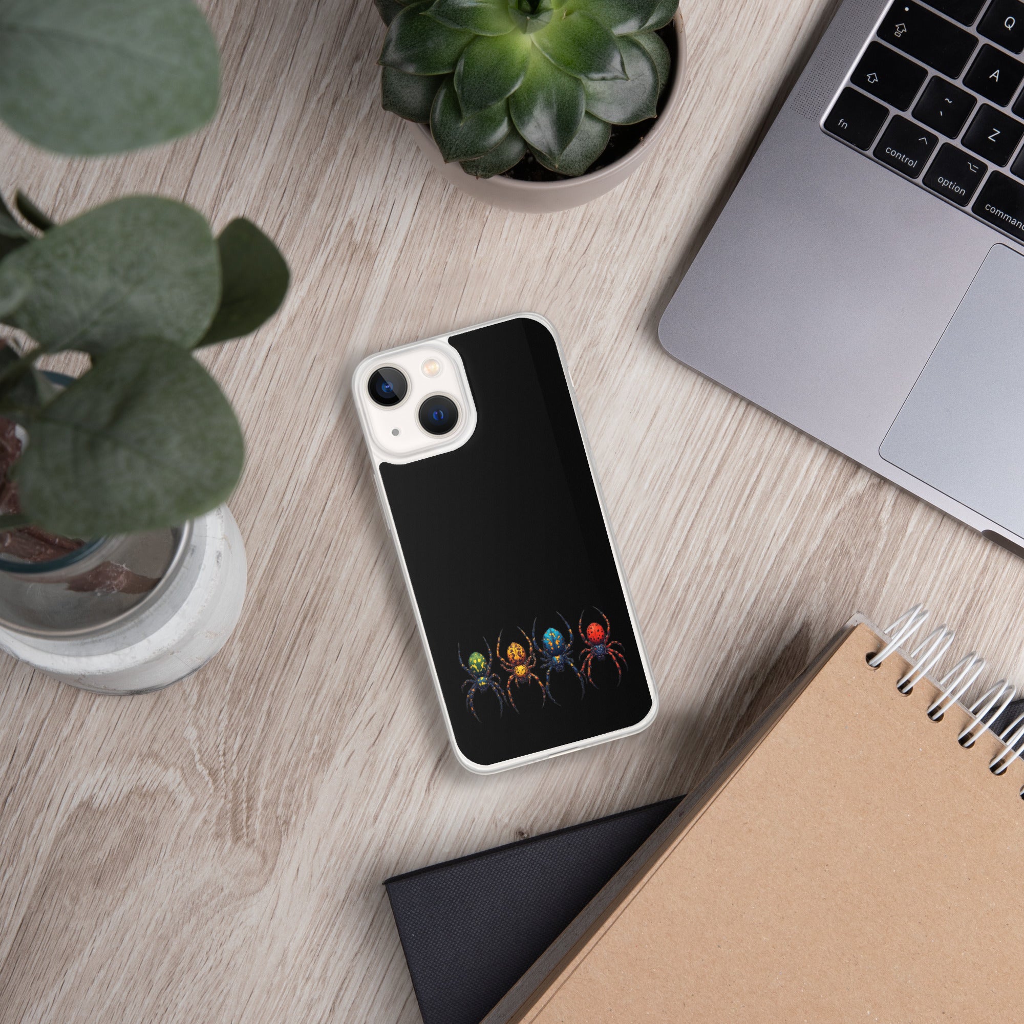 A black phone case with a graphic of four colorful spiders at the bottom, shown on a light wood desk with a laptop, plants, and notebooks.