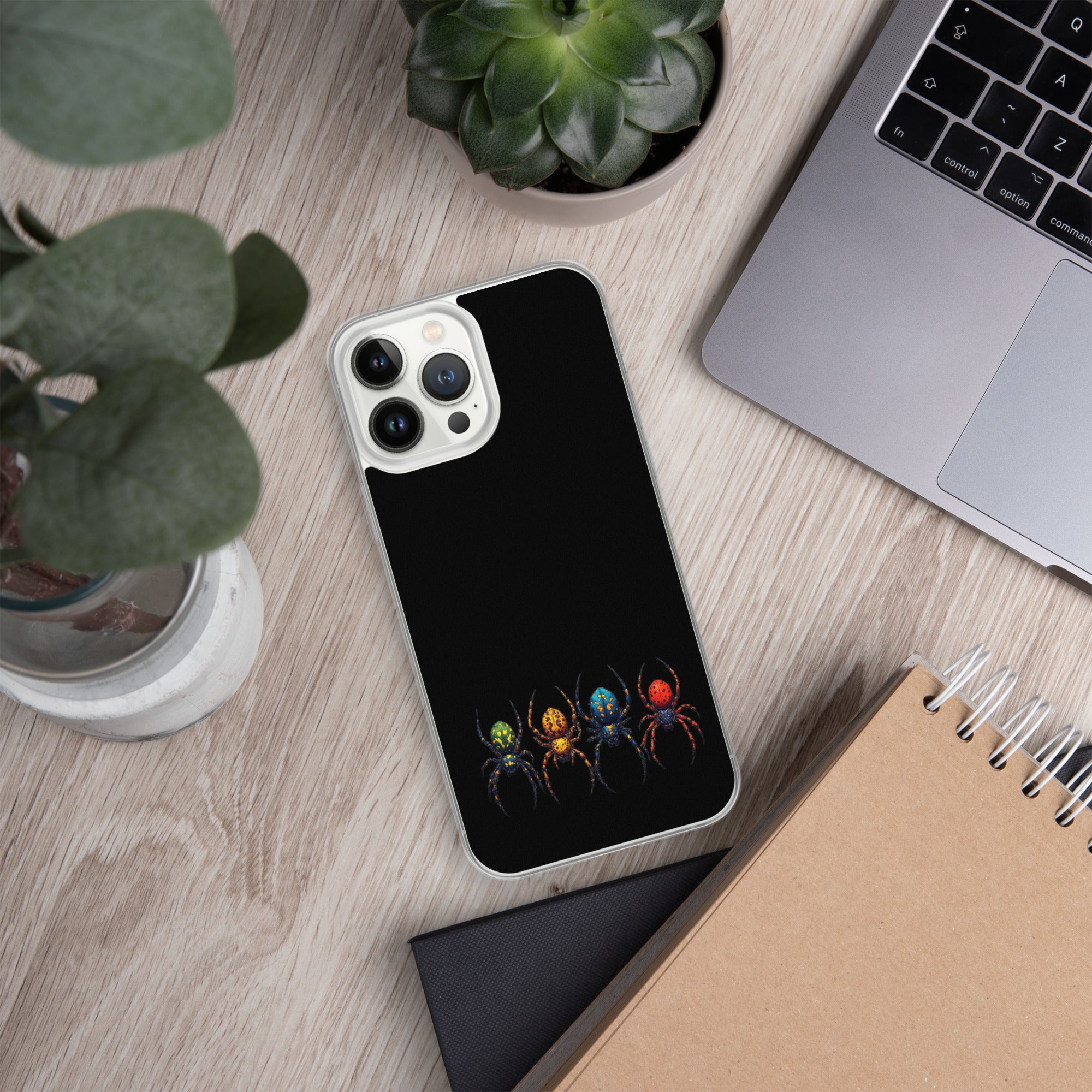 A black phone case with a colorful spider design, shown on a light wood desk with a laptop, plants, and notebooks.