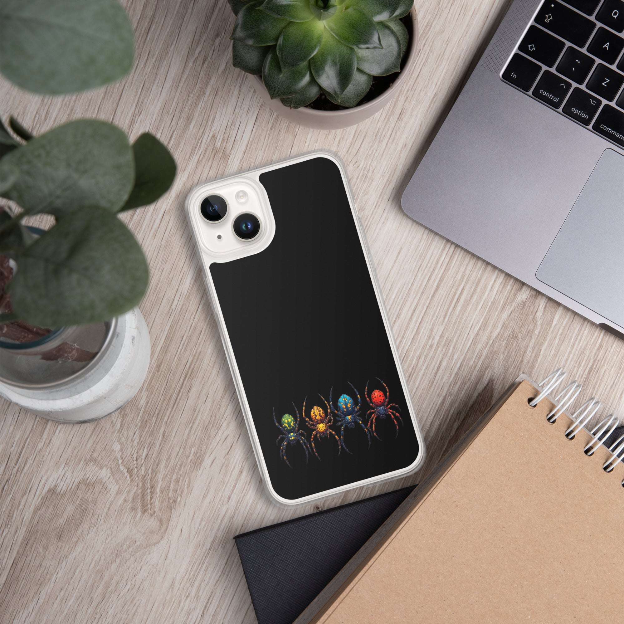 A black phone case with a colorful spider graphic, shown on a light wood desk surrounded by a laptop, plants, and a notebook.