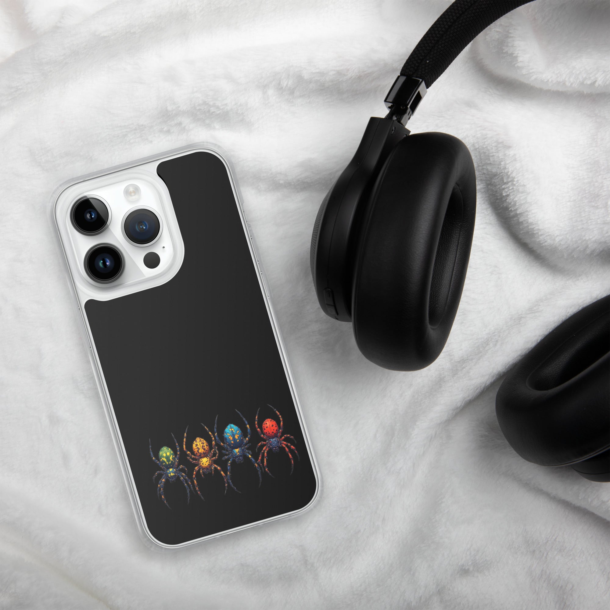 A clear-edged smartphone case with a black back panel and a colorful spider graphic, shown on a white textured blanket next to black headphones.