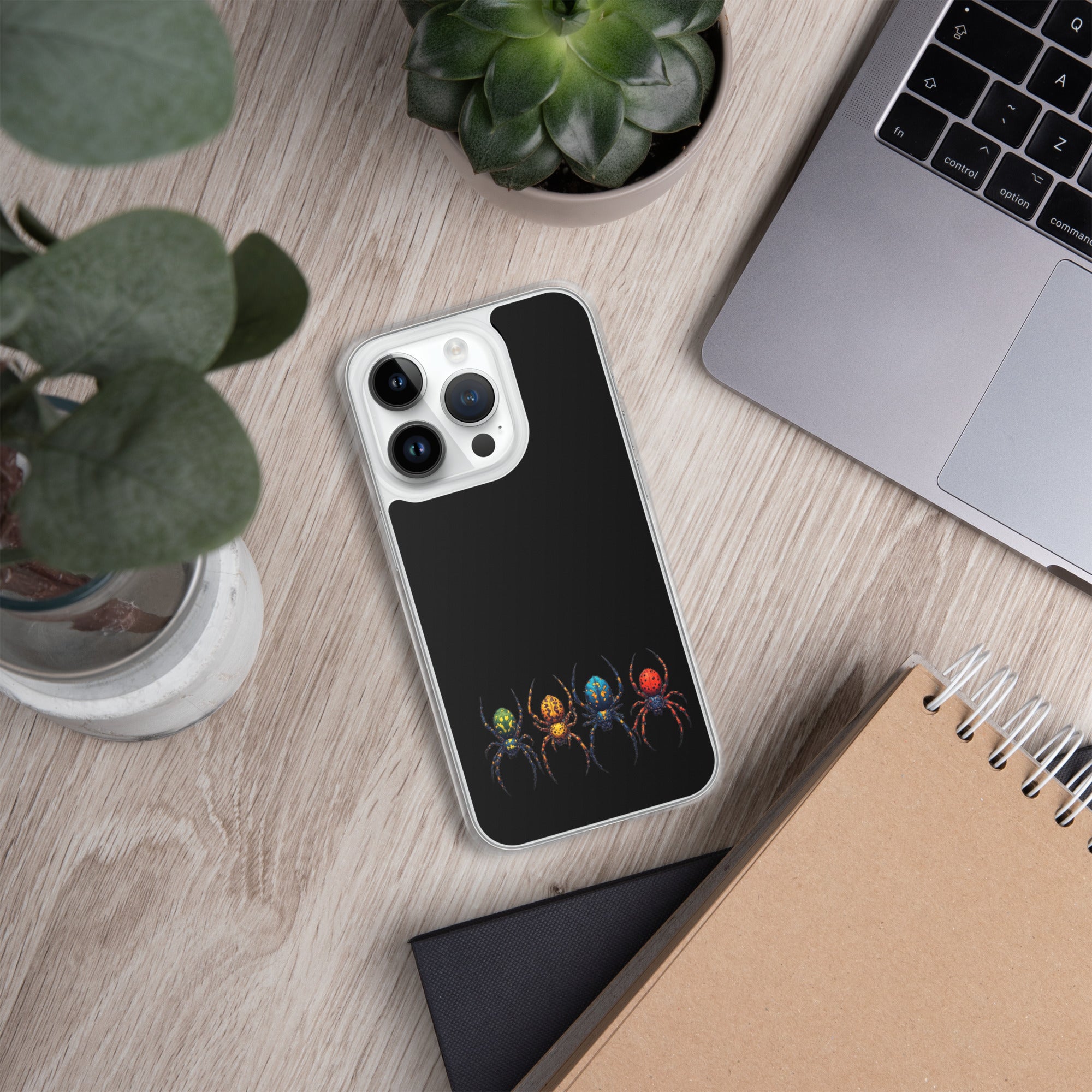 A black phone case with a colorful spider design, shown on a silver smartphone in a styled flat-lay setting with a laptop, plants, and notebooks on a wooden desk.