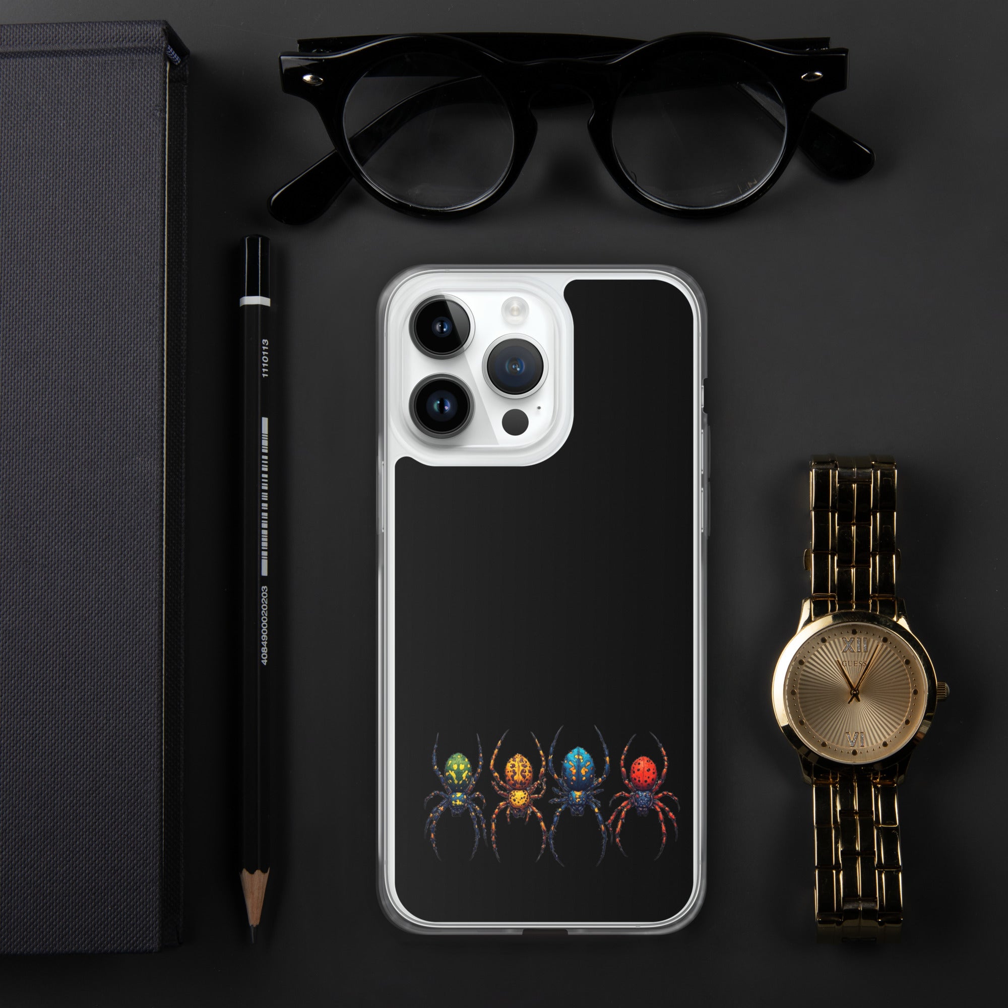 A clear iPhone case with a black back panel featuring a colorful graphic of four stylized spiders at the bottom, styled in a flat-lay with black eyeglasses, a black pencil, a dark textured box, and a gold-tone watch on a dark gray background.