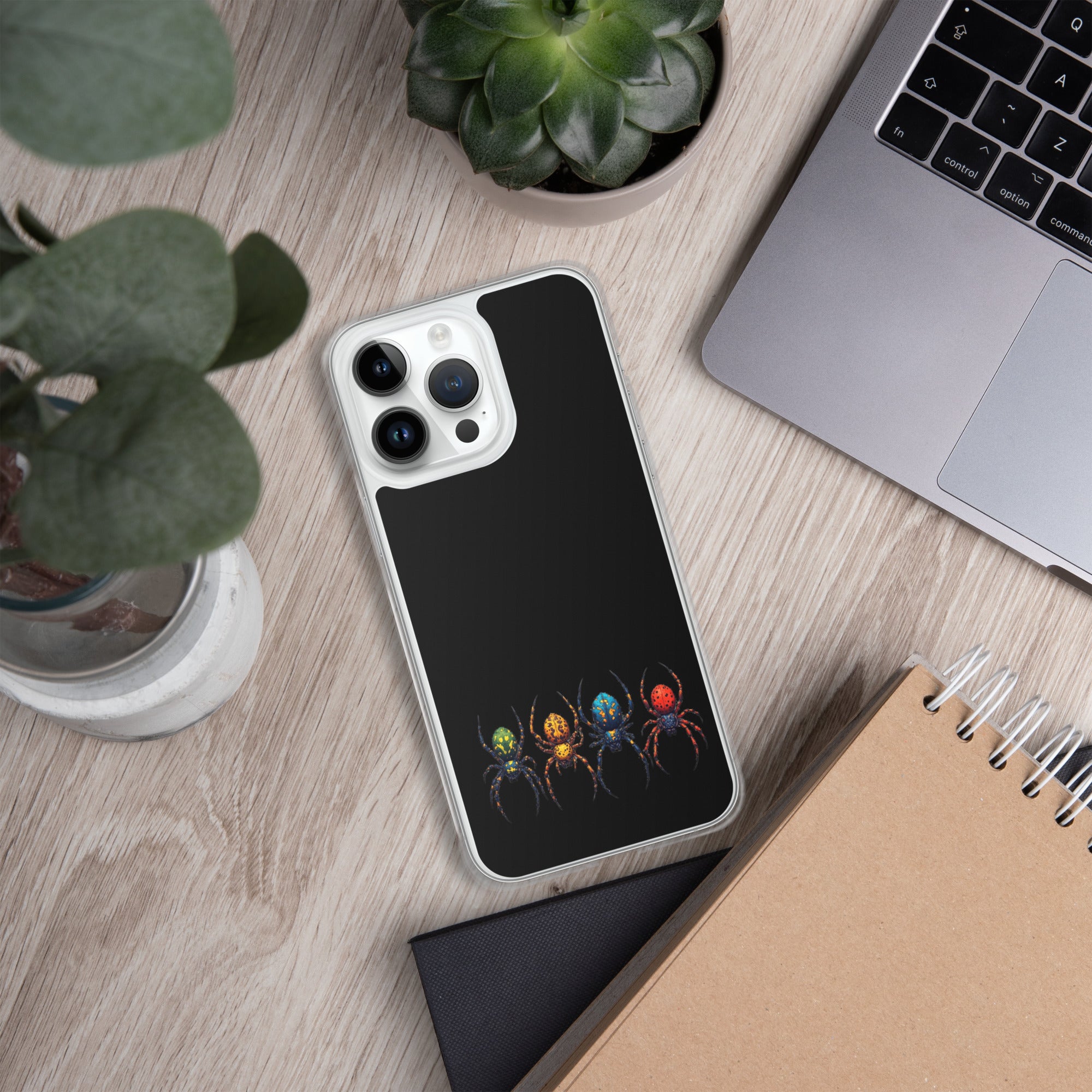 A black phone case with a colorful spider graphic, shown on a silver smartphone in a styled flat-lay desk setting with a laptop, plants, and notebooks.