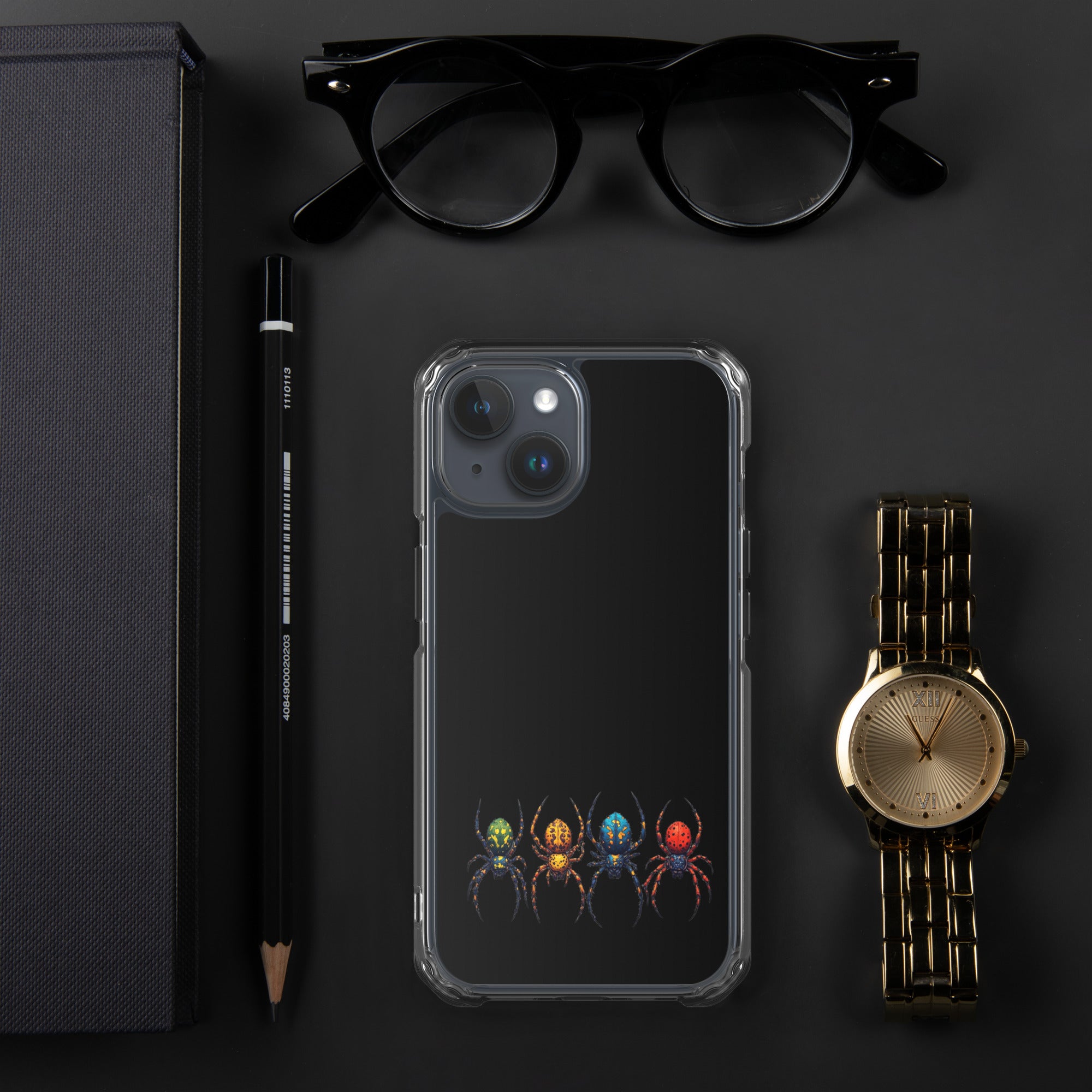 A clear iPhone case with a graphic of four colorful spiders at the bottom, displayed in a flat-lay arrangement with black eyeglasses, a black pencil, a black notebook, and a gold watch on a dark gray background.