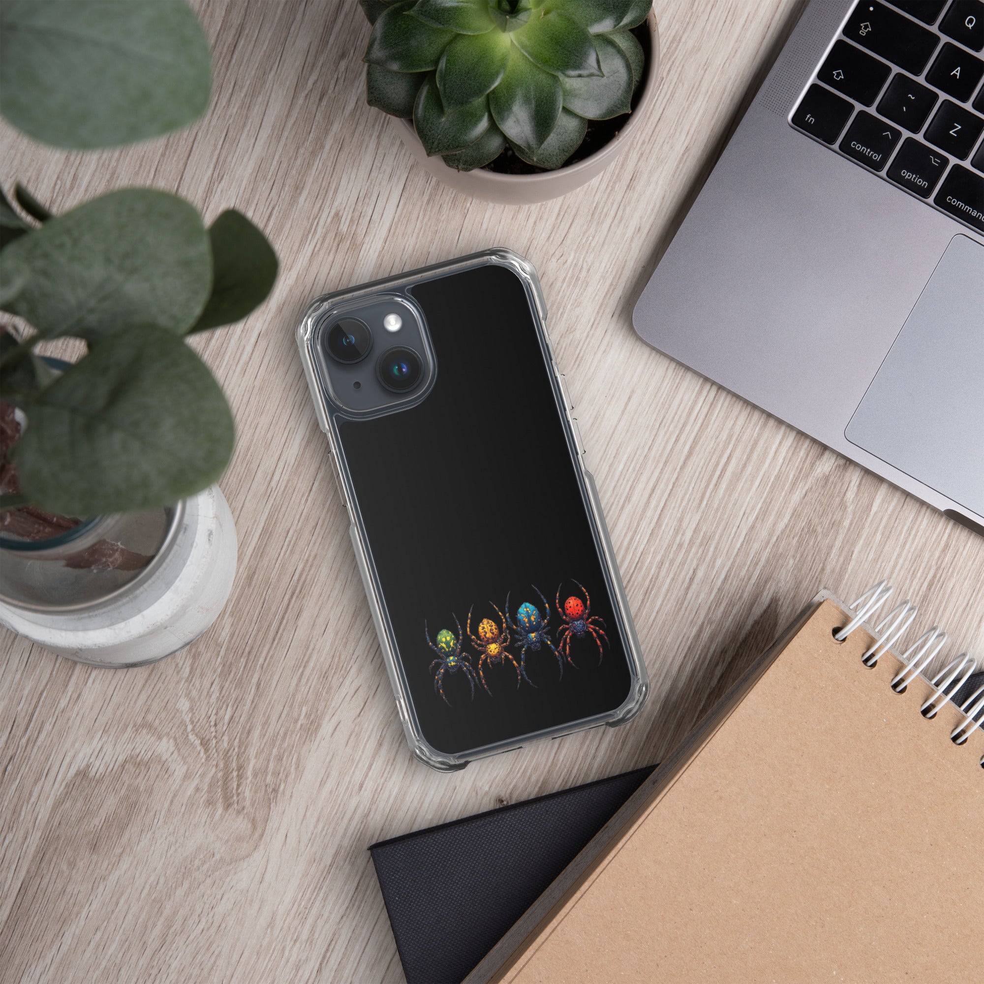 A clear phone case with a black back panel featuring a graphic of four colorful spiders, shown in a styled flat-lay on a light wood desk next to a laptop, notebook, and plants.