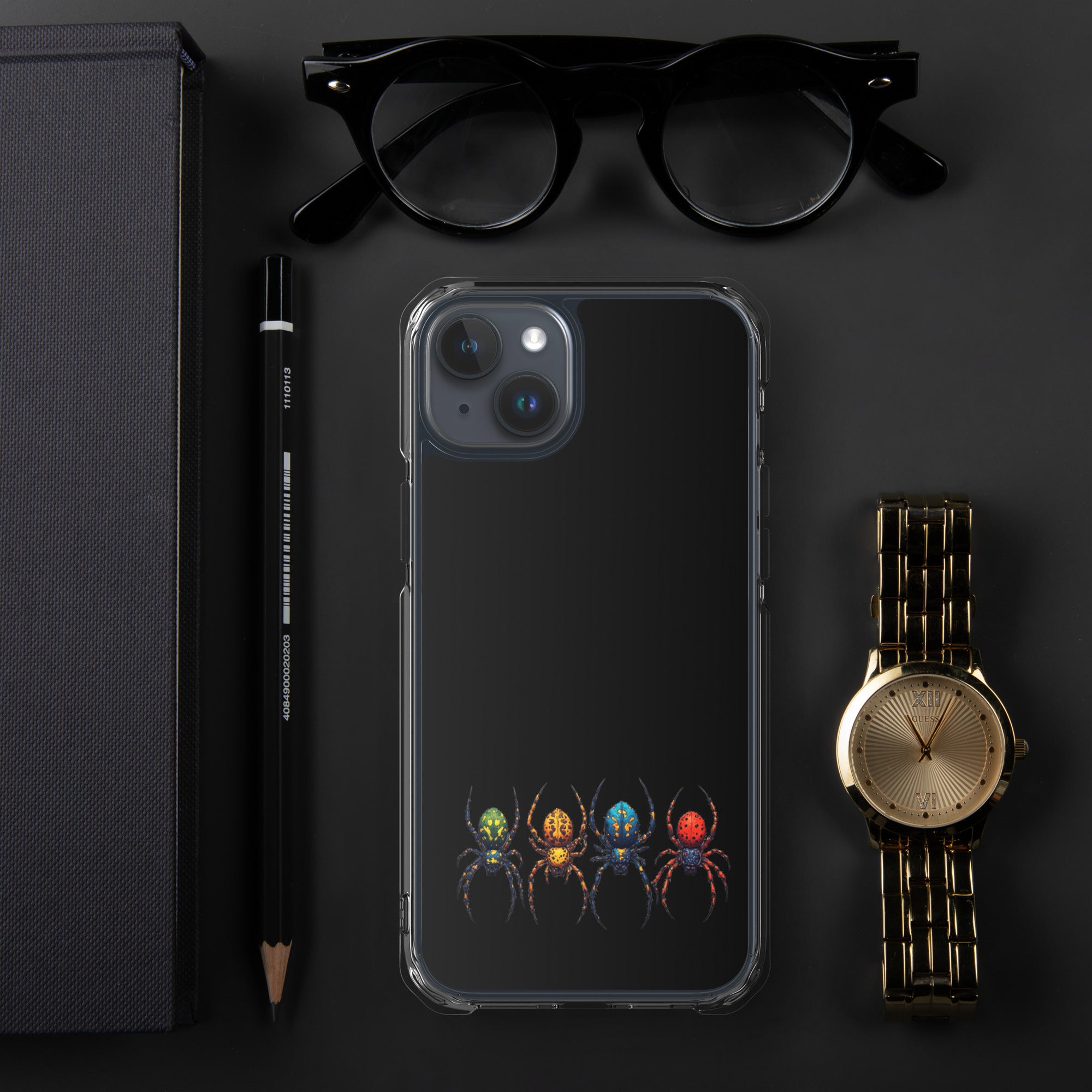 A clear smartphone case with a printed graphic of four colorful spiders at the bottom, displayed in a flat-lay arrangement on a dark surface alongside black eyeglasses, a black pencil, a gold-toned watch, and a dark textured box.