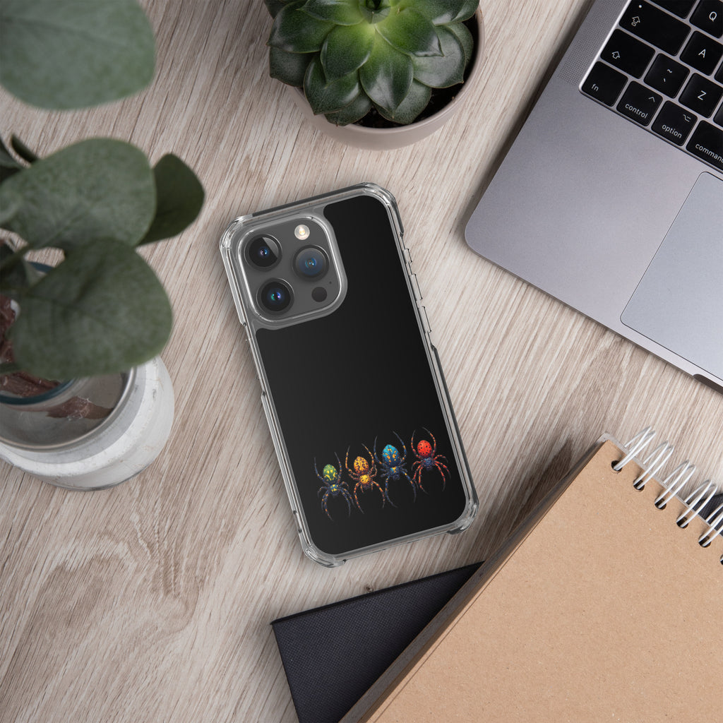 A clear phone case with a black back panel featuring a graphic of four colorful spiders, shown in a styled flat-lay desk setting with a laptop, notebook, and plants.
