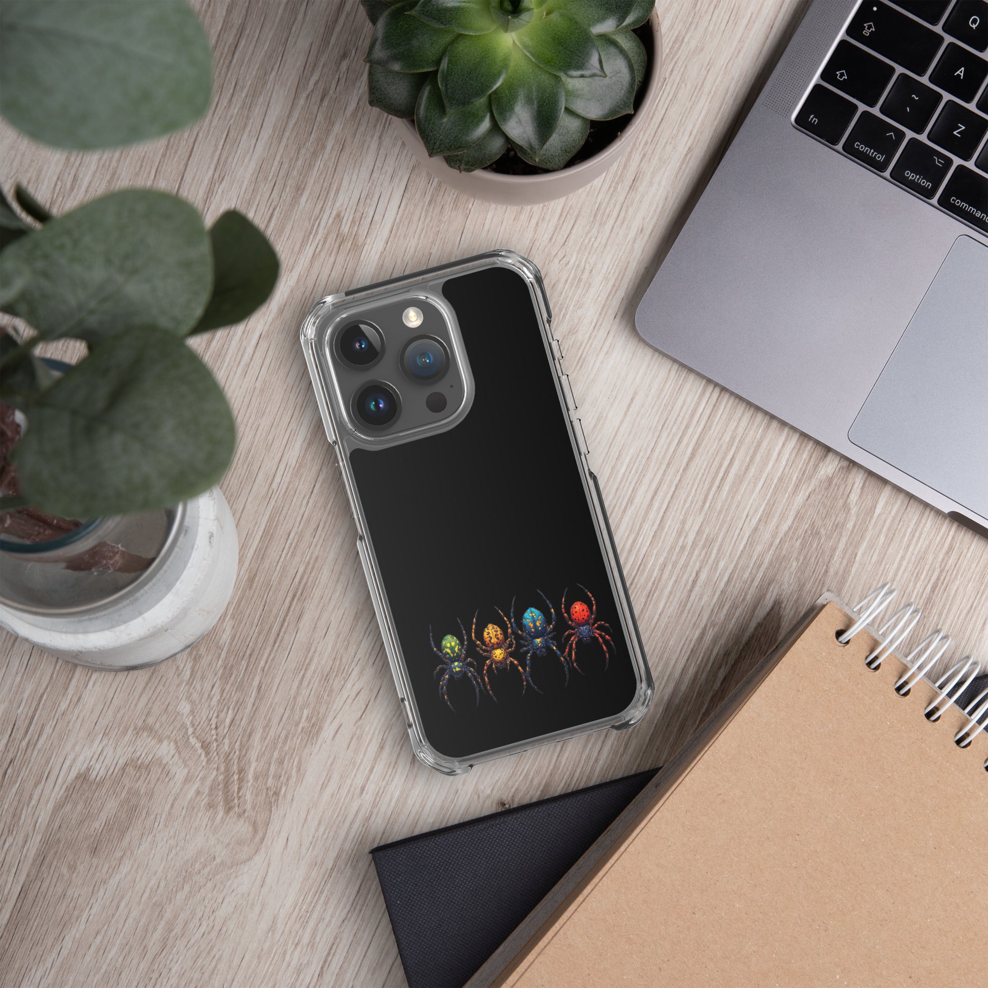 A clear phone case with a black back panel featuring a graphic of four colorful spiders, shown in a styled flat-lay desk setting with a laptop, notebook, and plants.