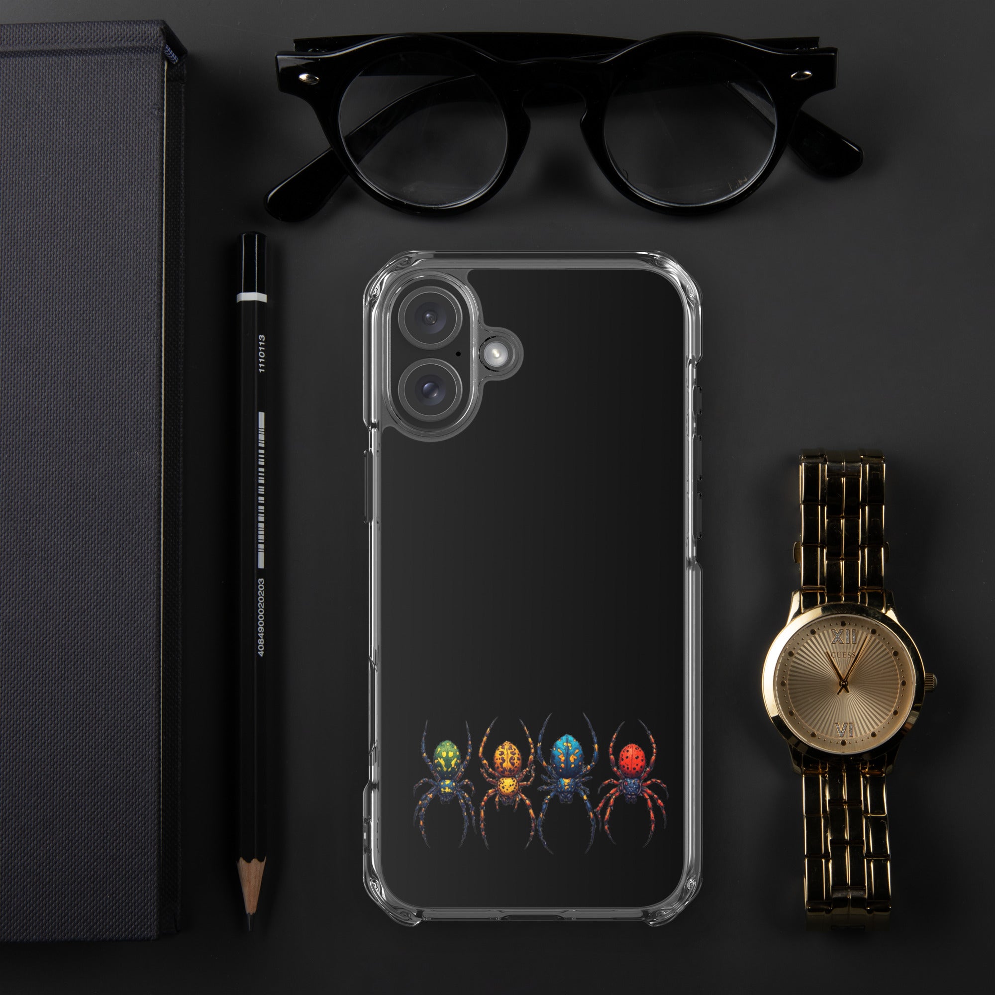 A clear protective phone case with a black back panel featuring a colorful graphic of four stylized spiders at the bottom, displayed in a flat-lay arrangement with black eyeglasses, a black pencil, a dark textured box, and a gold-tone wristwatch on a dark gray background.
