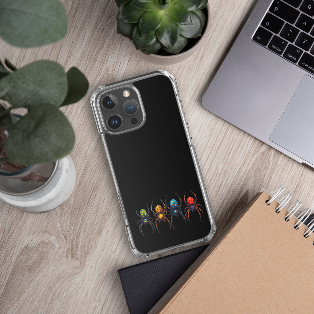 A clear-edged phone case with a black back panel featuring a colorful graphic of four stylized spiders, shown on a wooden desk next to a laptop, notebook, and plants.