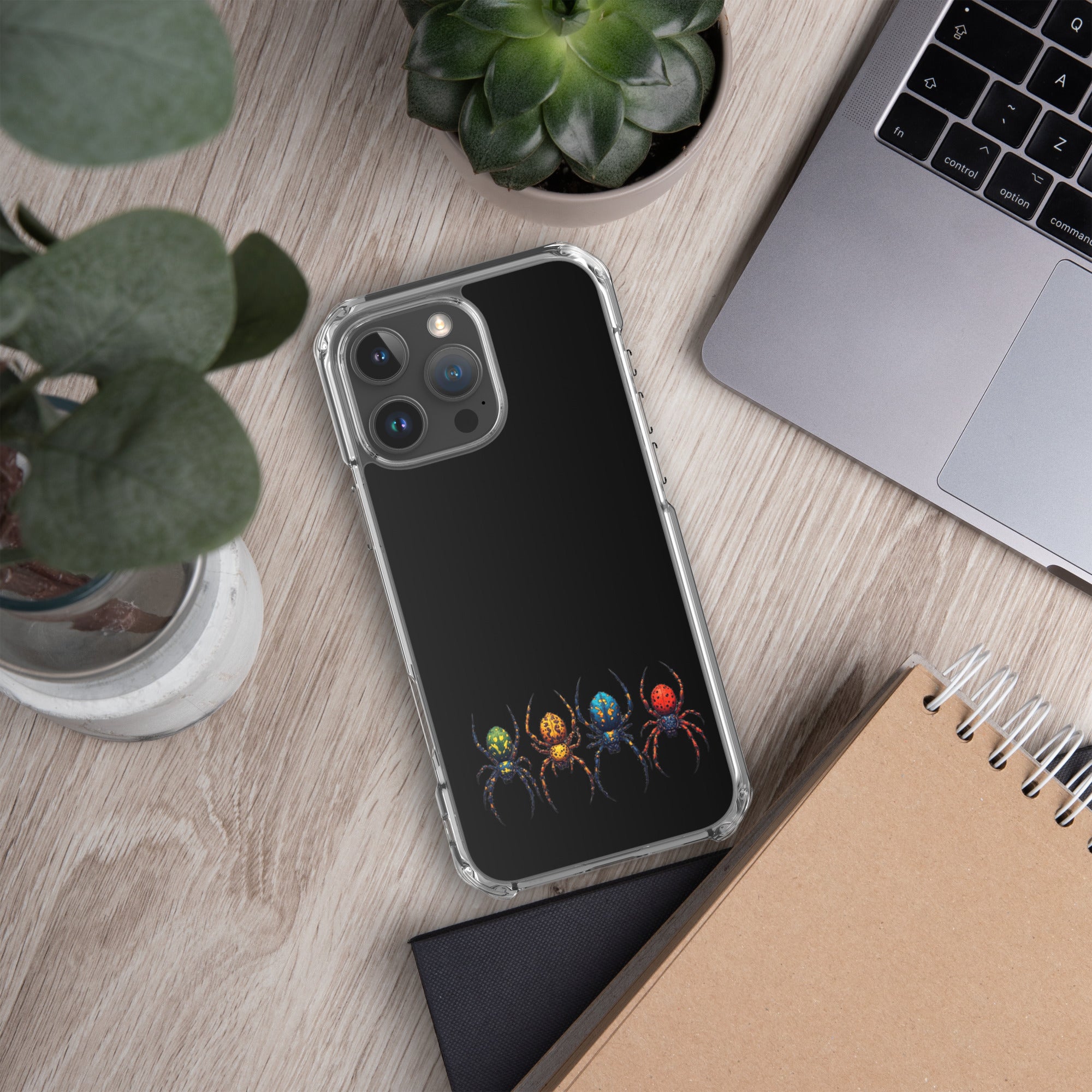 A clear-edged phone case with a black back panel featuring a colorful graphic of four stylized spiders, shown on a wooden desk next to a laptop, notebook, and plants.