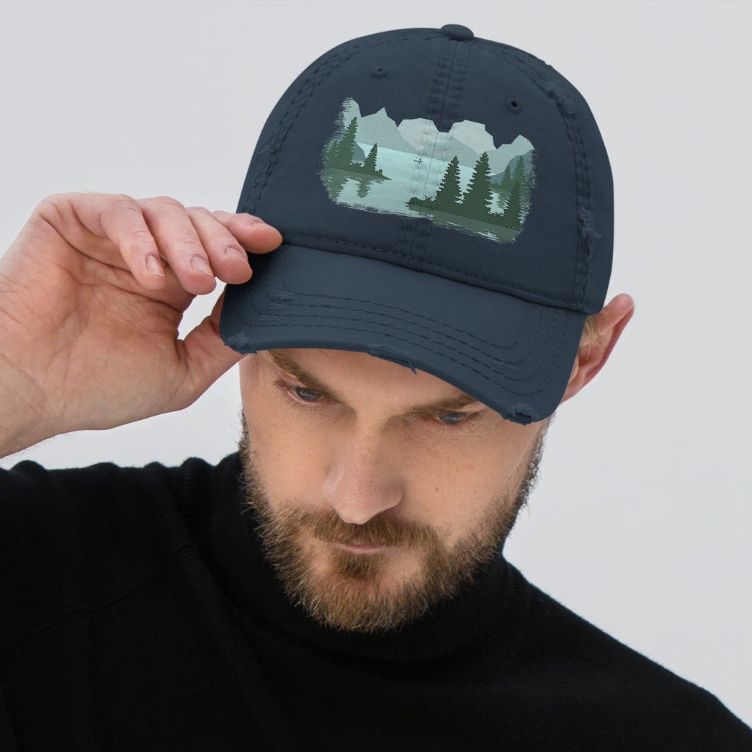 Man adjusting the bill of a navy blue distressed graphic dad hat featuring a fisherman boat lake scene print, relaxed fit fishing cap with adjustable back closure. 