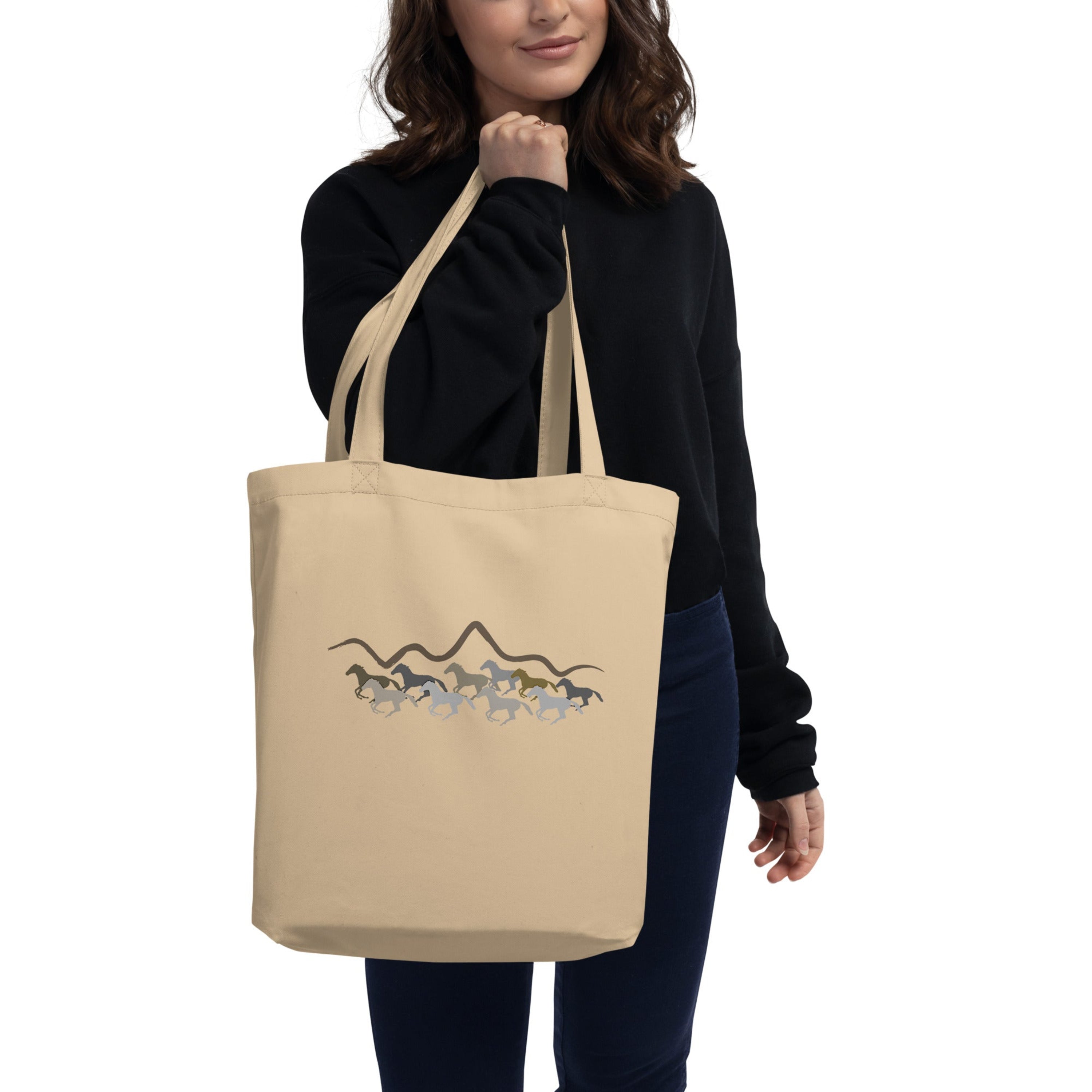 A woman holding a tote bag with a running wild horses and mountain silhouette graphic design in earth tones.