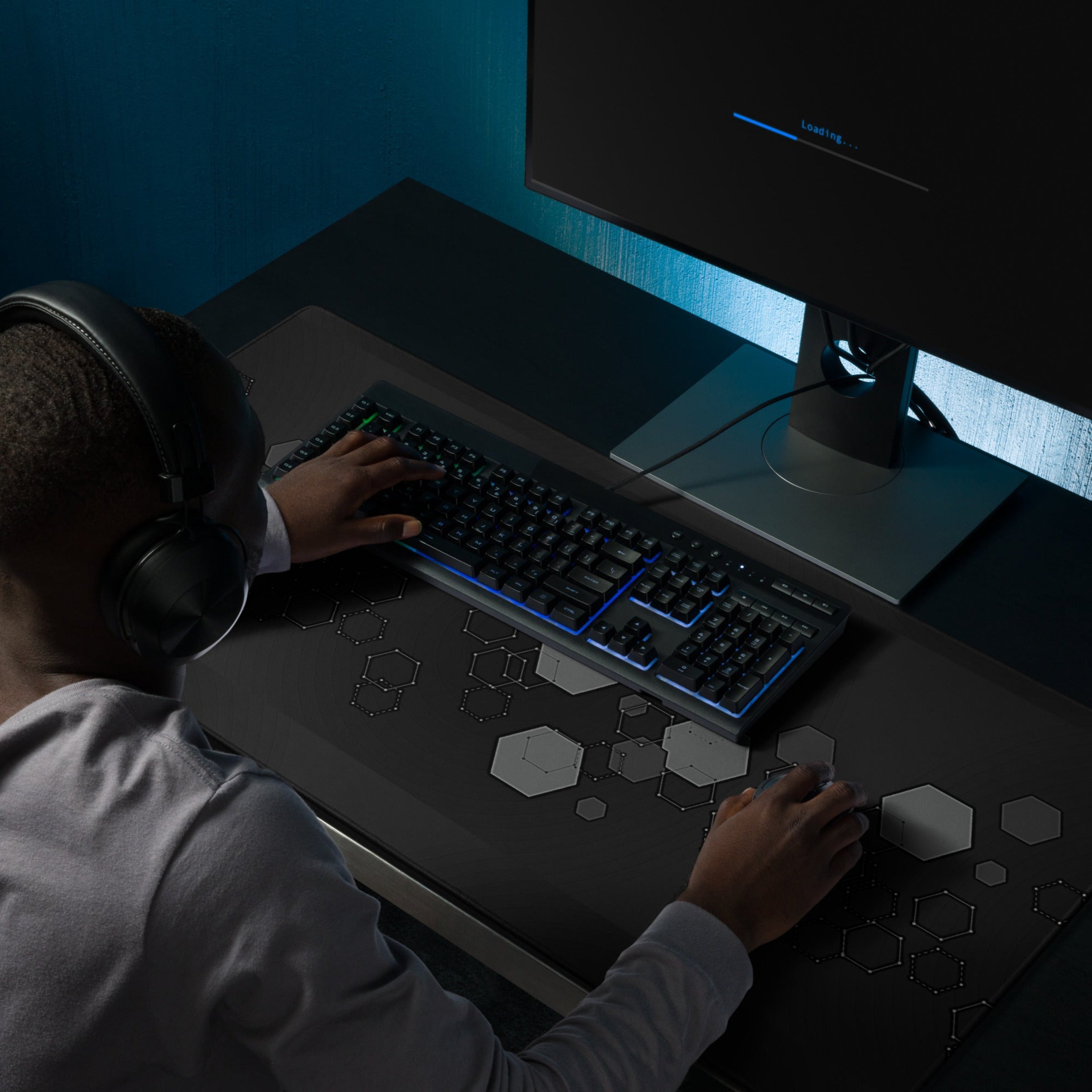 Person wearing headphones using a computer with a Dark extended gaming desk mat with grey hexagon geometric pattern. 