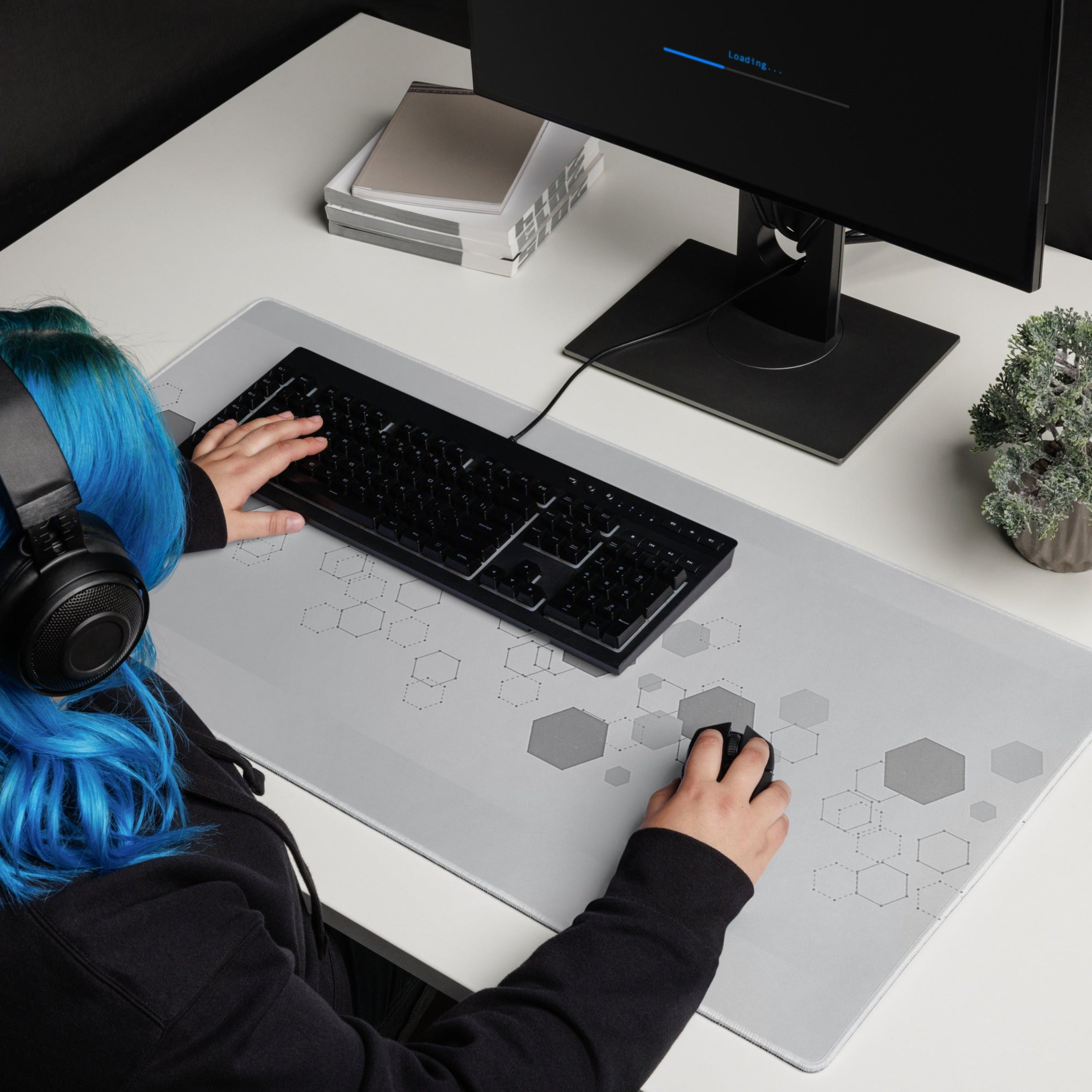Person with blue hair using a computer at a desk with a keyboard and mouse on a extended gamer mouse pad with a hexagon graphic design.