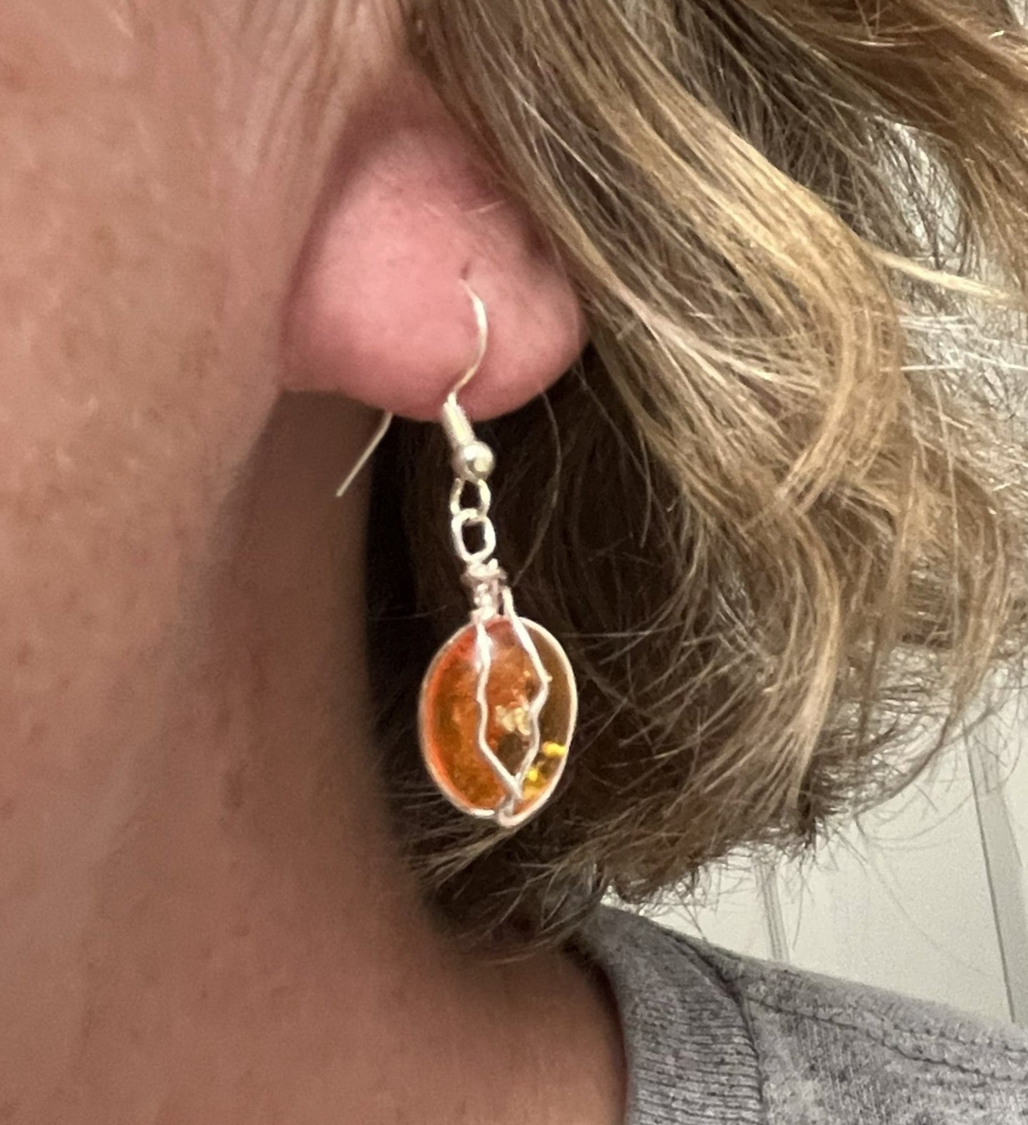 A close-up of a person's ear wearing a dangling earring with a silver-toned fishhook and an oval amber-orange stone in a silver setting.