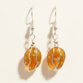 A pair of handmade dangle earrings featuring oval amber-colored glass beads wrapped in silver wire, hanging from silver fishhook ear wires on a brown textured cardboard background.